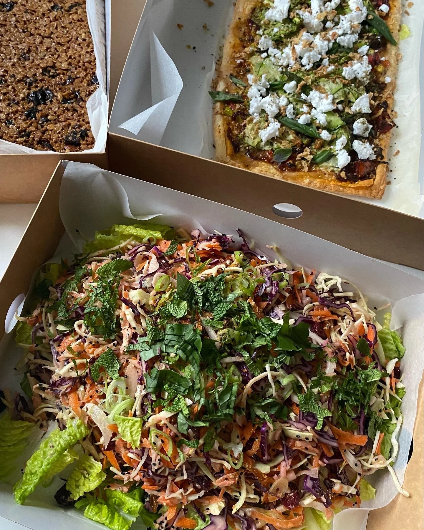 Boxed salad topped with mixed greens and shredded vegetables, a rectangular flatbread with green herbs, cheese, and toppings, and a square-baked dessert with nuts and berries.