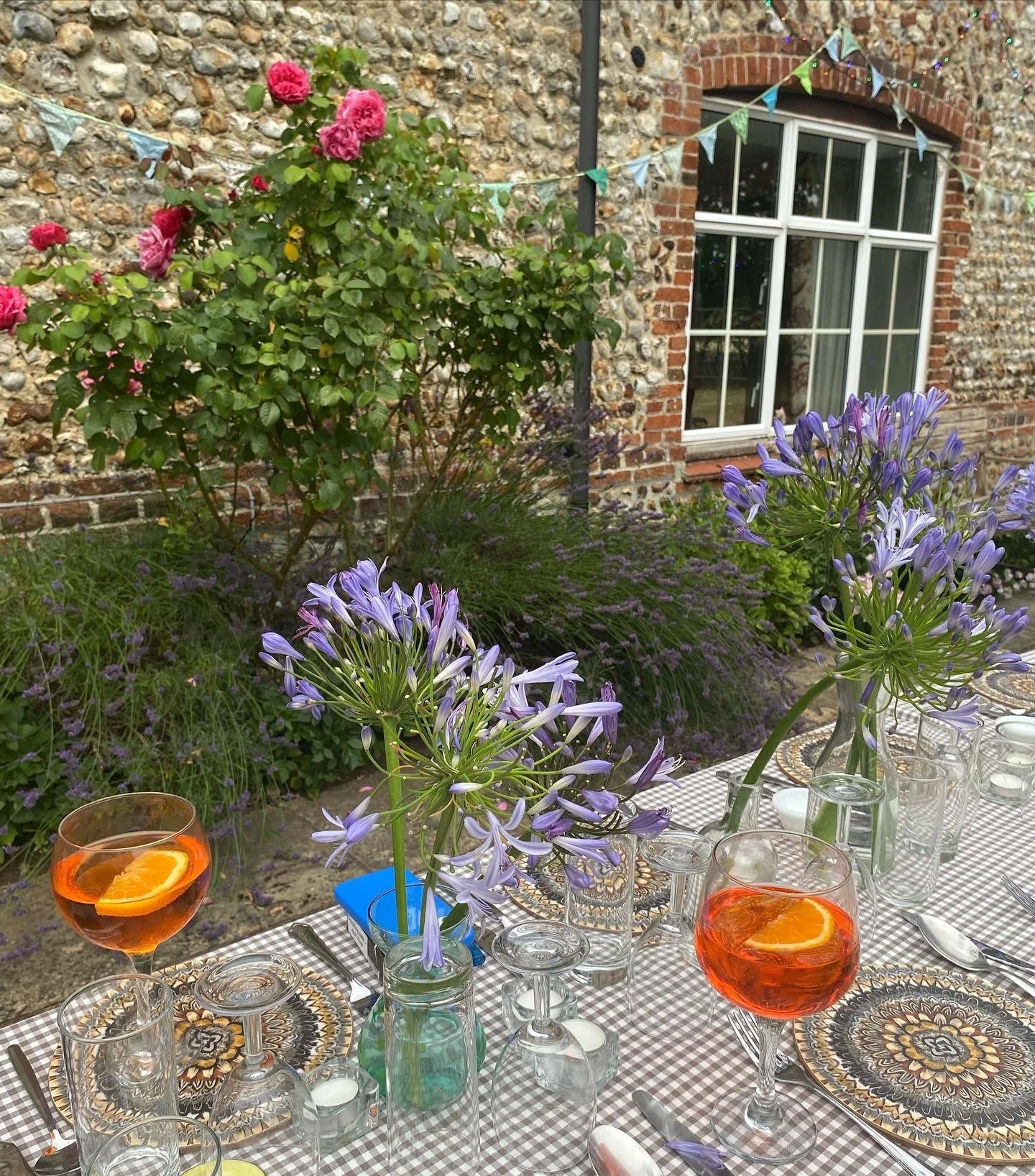 A garden party table setting with purple flowers in vases, glasses of orange-colored drinks with orange slices, set on a checkered tablecloth outside next to a brick and stone building with a window and bunting.