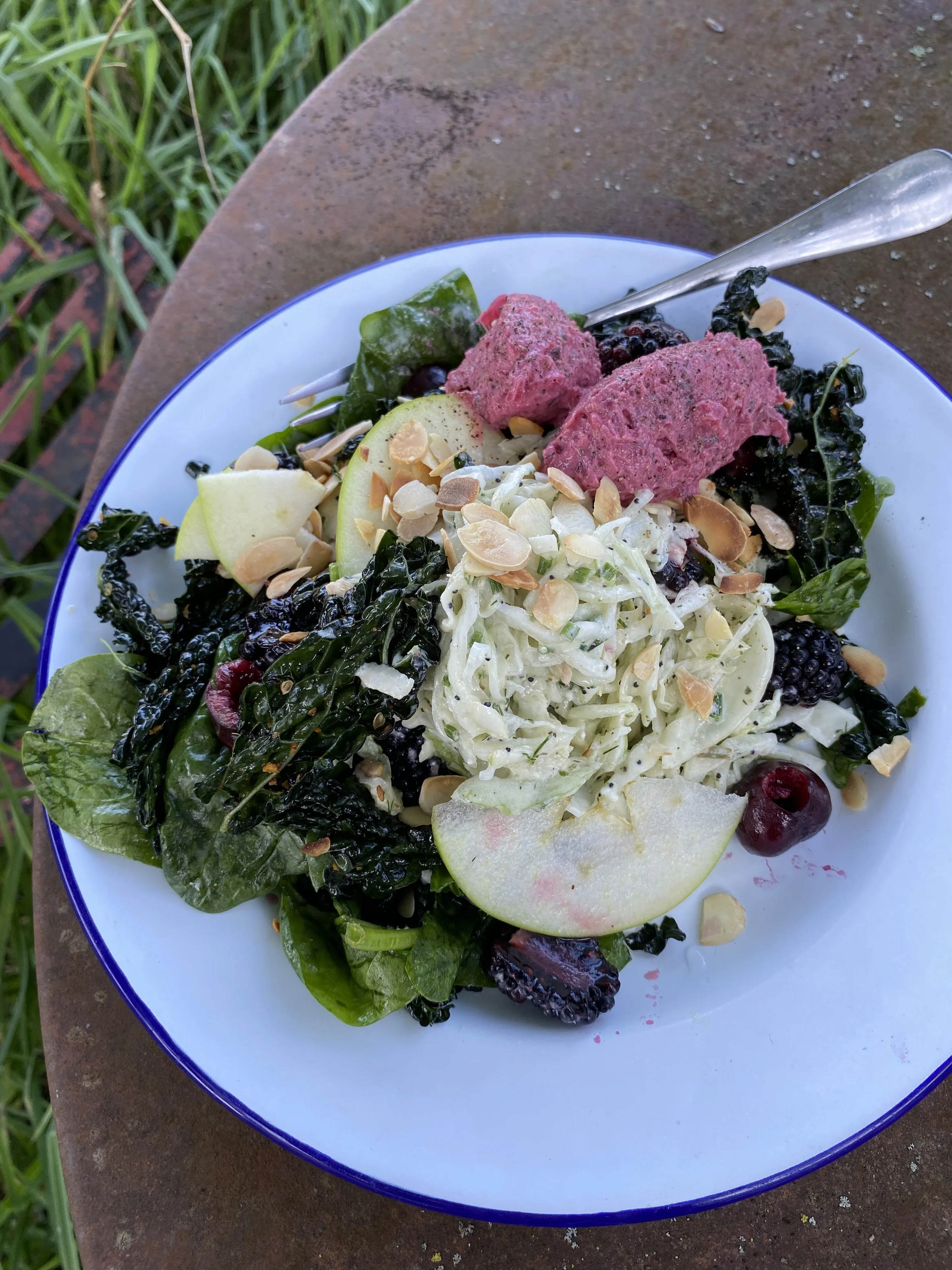 A colorful salad with greens, pear slices, berries, shredded cheese, slivered almonds, and two pink scoops of a berry dessert, served on a white plate with a blue rim