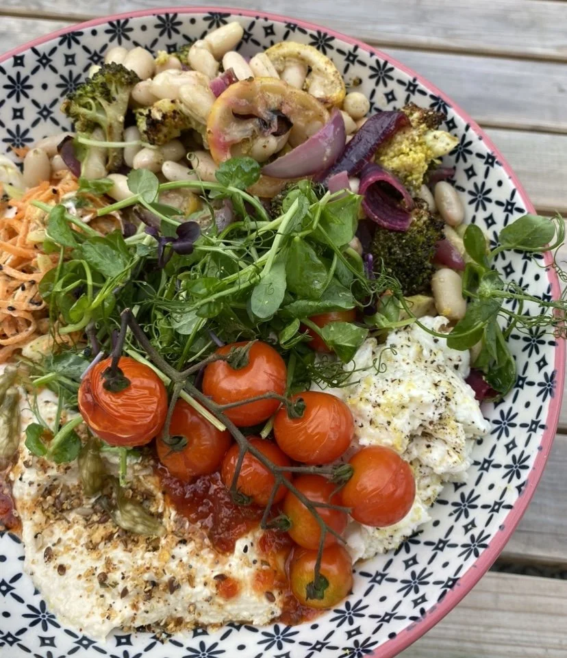 A bowl of fresh salad with cherry tomatoes, greens, roasted vegetables, grains, beans and a poached egg, on a wooden surface.