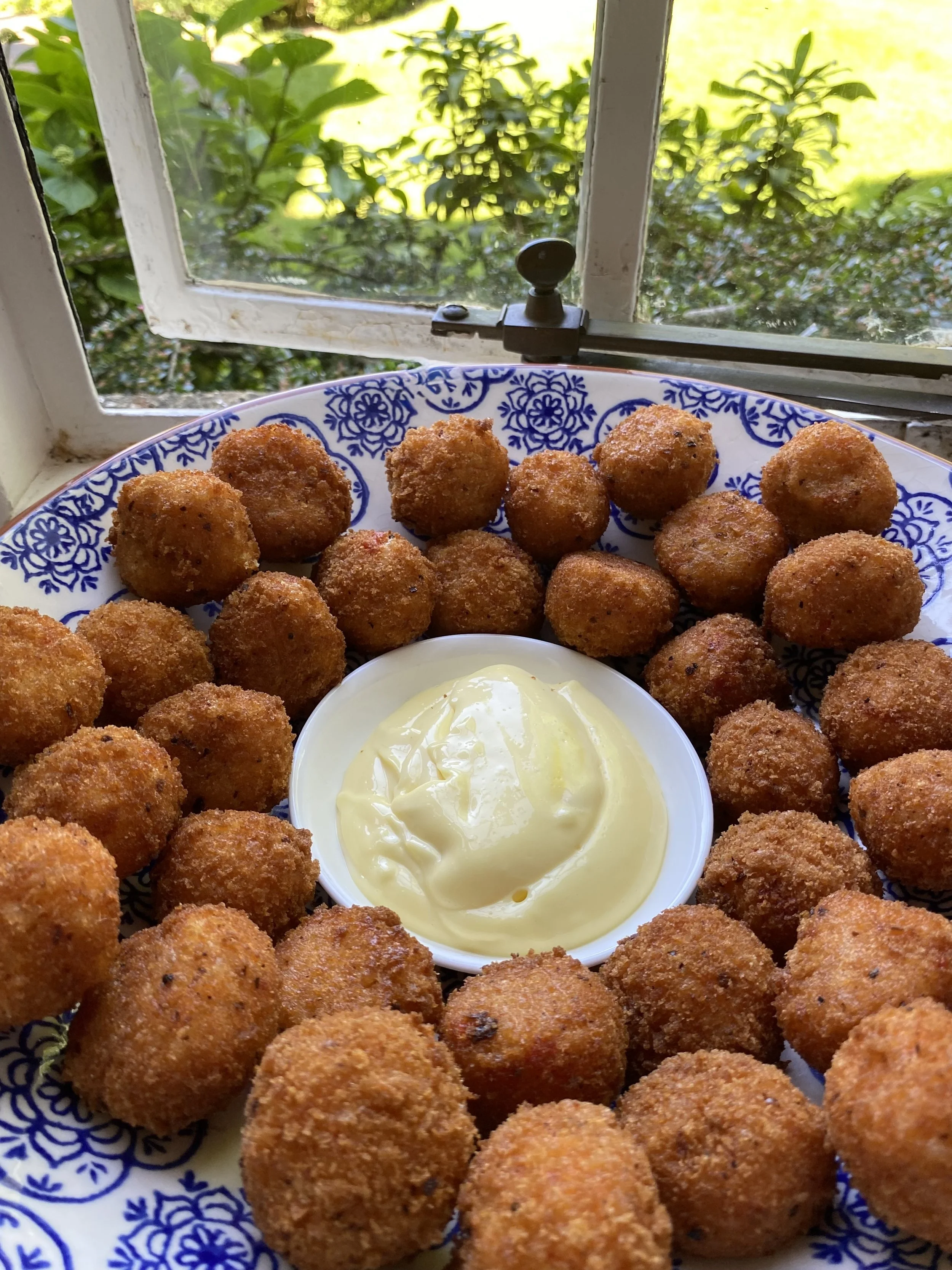 A plate of fried arancini appetizers, arranged in a circular pattern around a small bowl of creamy aioli, set on a decorative blue and white plate near a window with greenery outside.