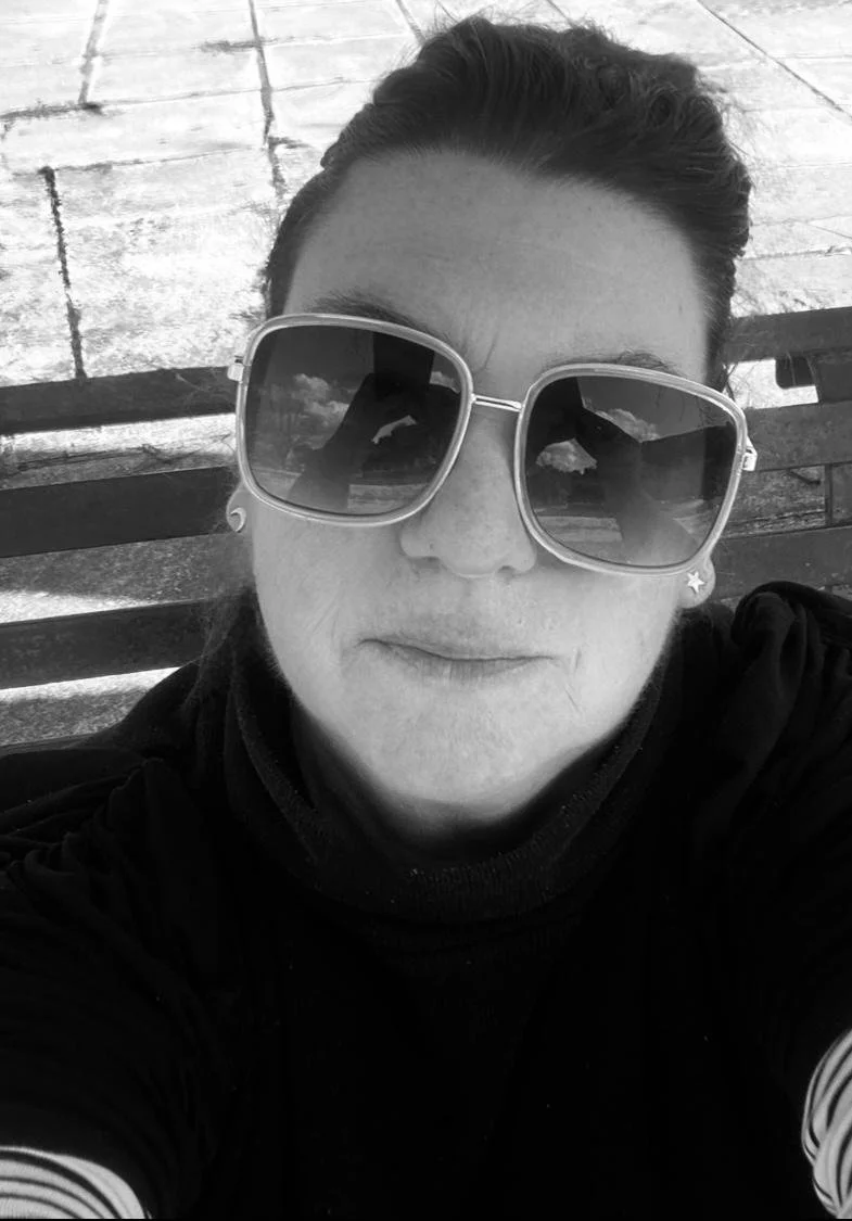 A woman with tied-back hair wearing large sunglasses, sitting on a bench outdoors in black and white.