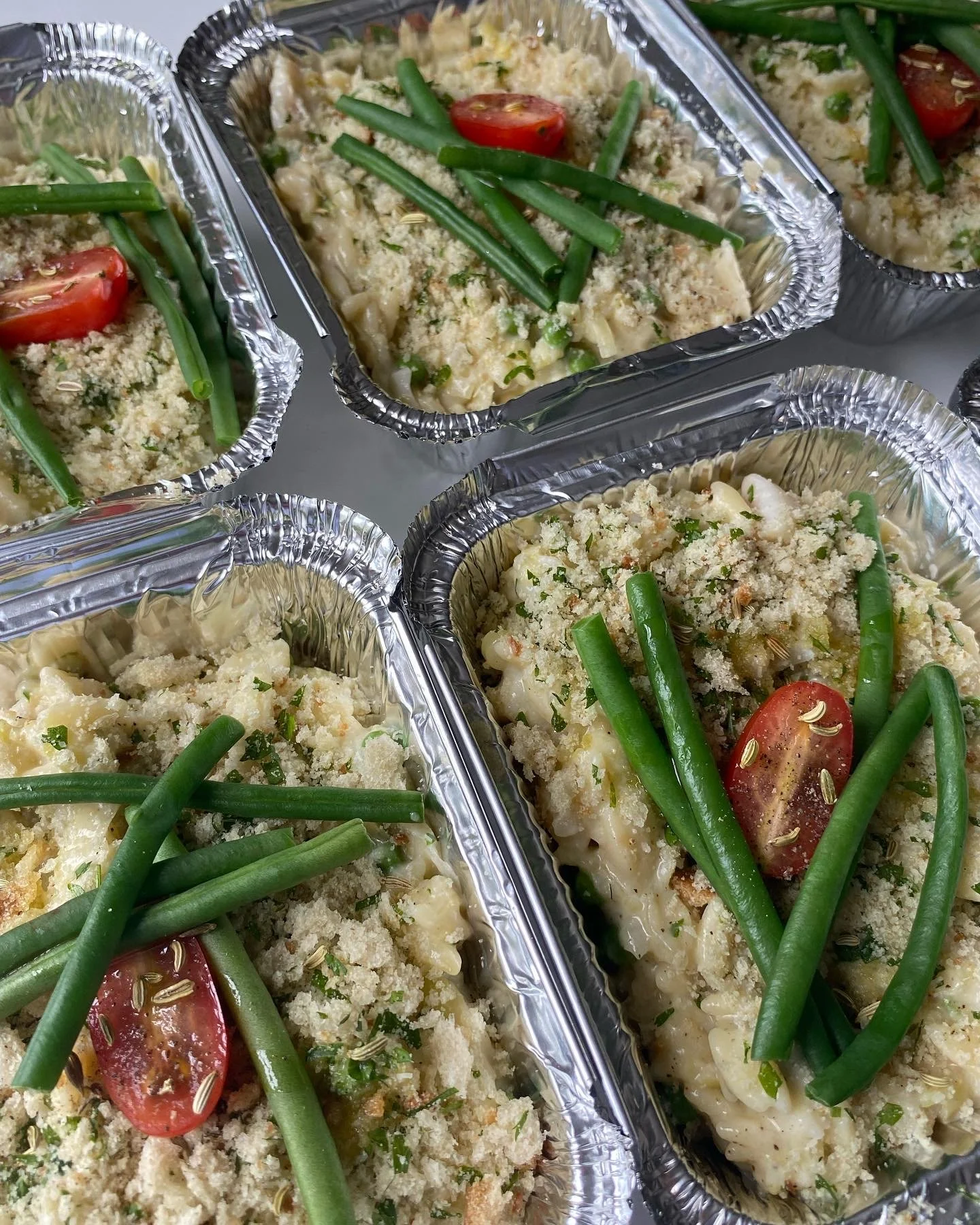 Multiple aluminum foil containers filled with baked pasta topped with green beans, cherry tomato slices, grated cheese, and herbs.