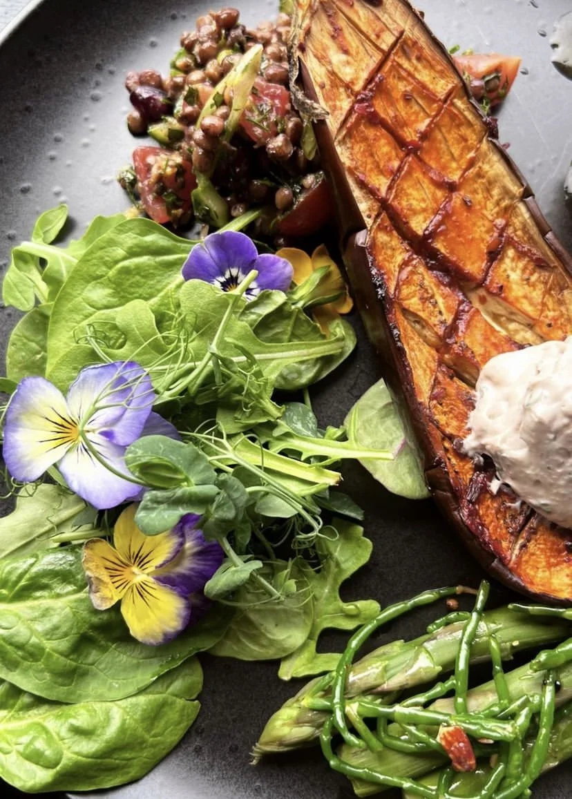 Grilled butternut squash with colourful salad, fresh greens with edible flowers, and cooked green beans on a black plate.
