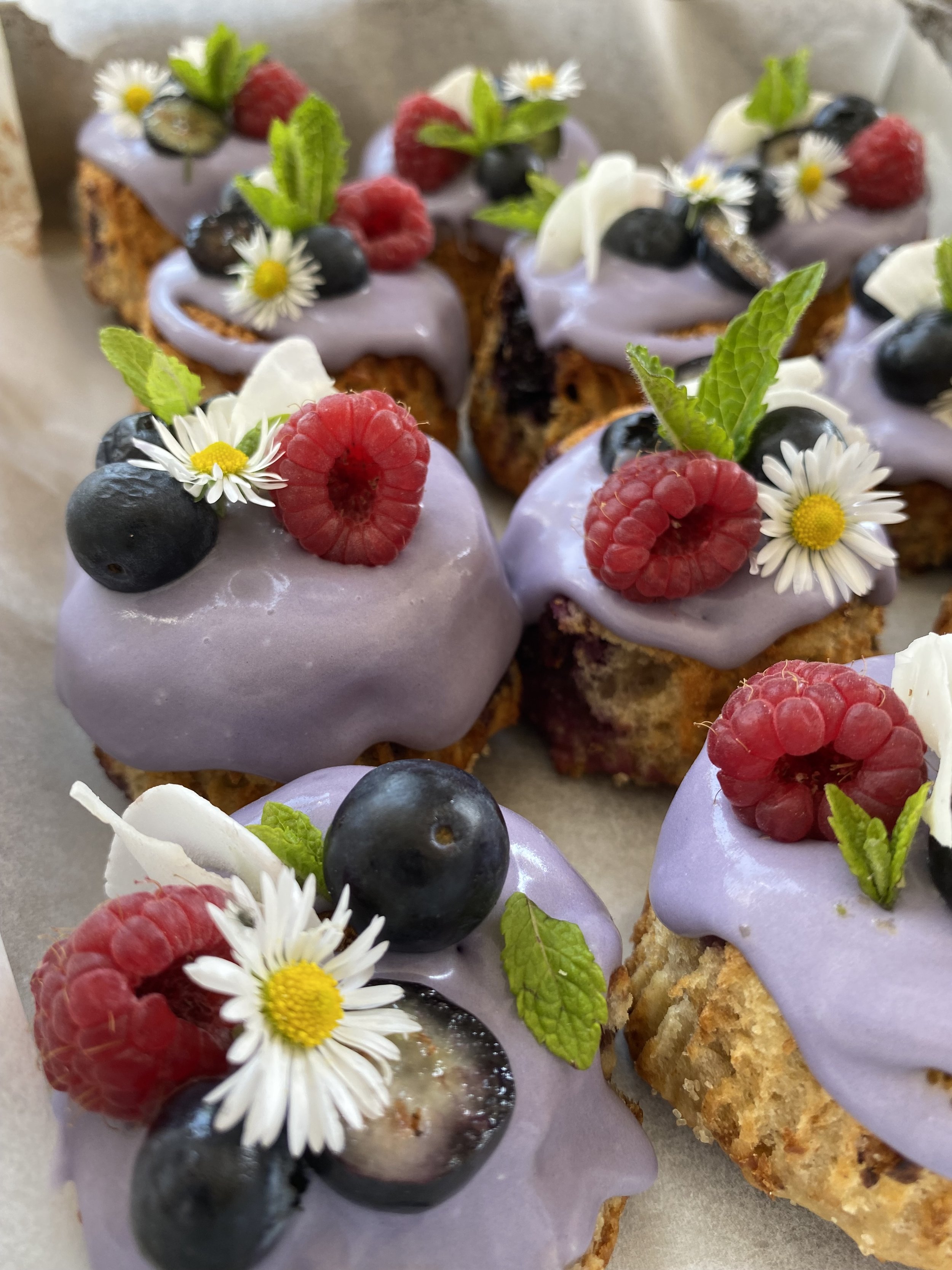 Mini cakes topped with purple icing, fresh blackberries, raspberries, mint leaves, and small white daisies.
