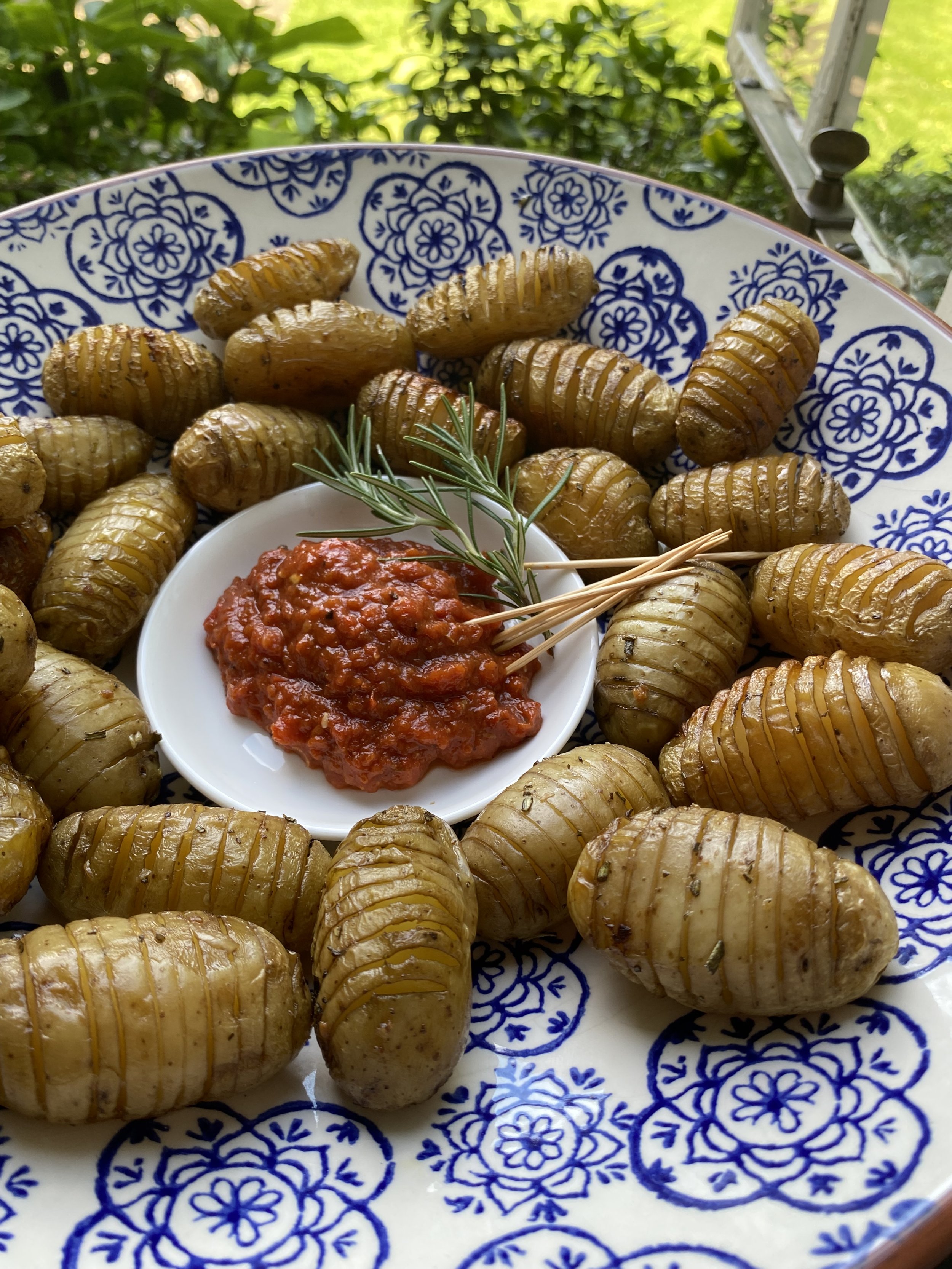 Roasted Hasselback potatoes served with a side of tomato-based dipping sauce garnished with a sprig of rosemary, on a decorative blue and white platter.
