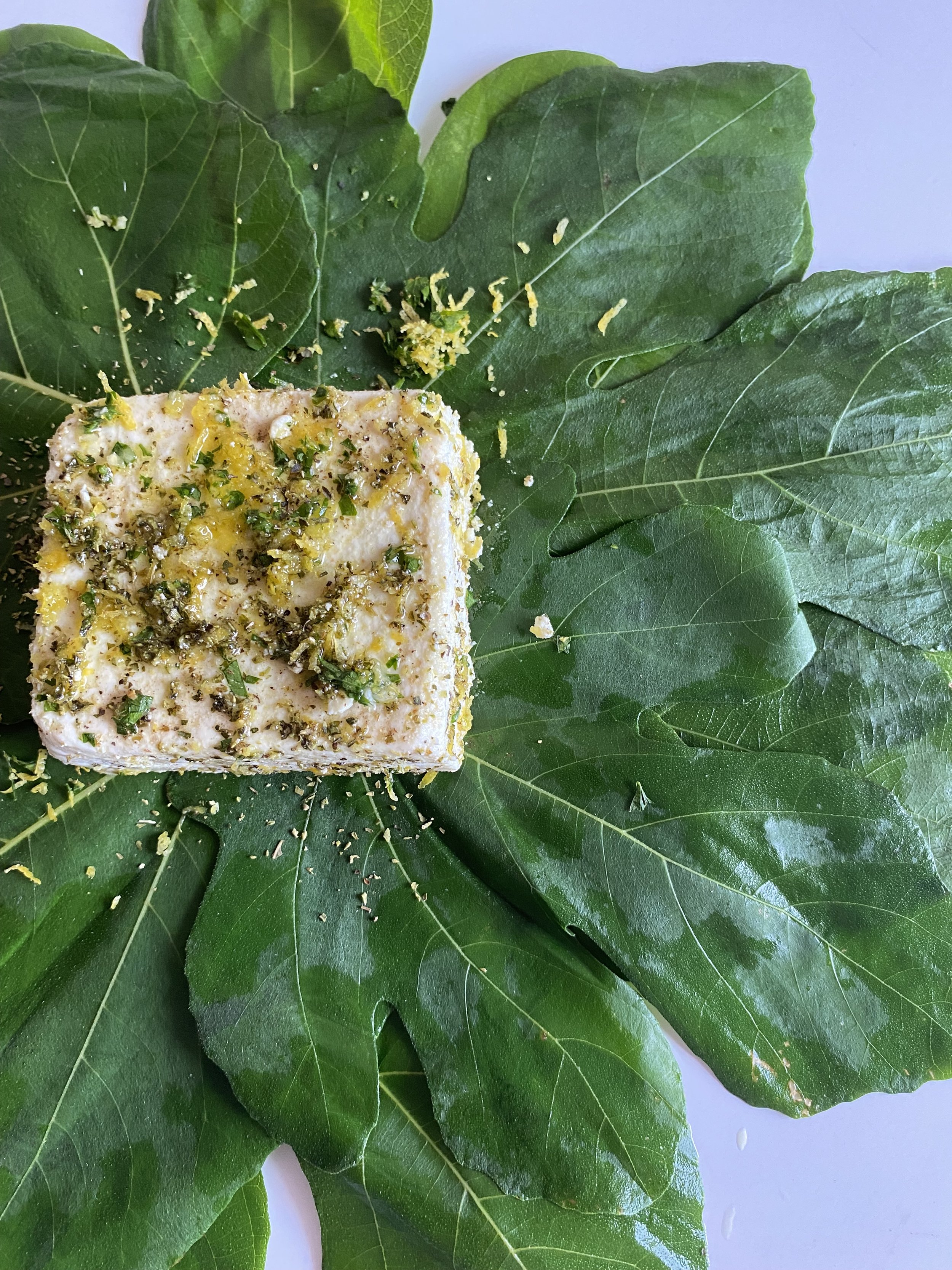 A piece of seasoned fish topped with herbs and lemon zest, placed on large green fig leaves.