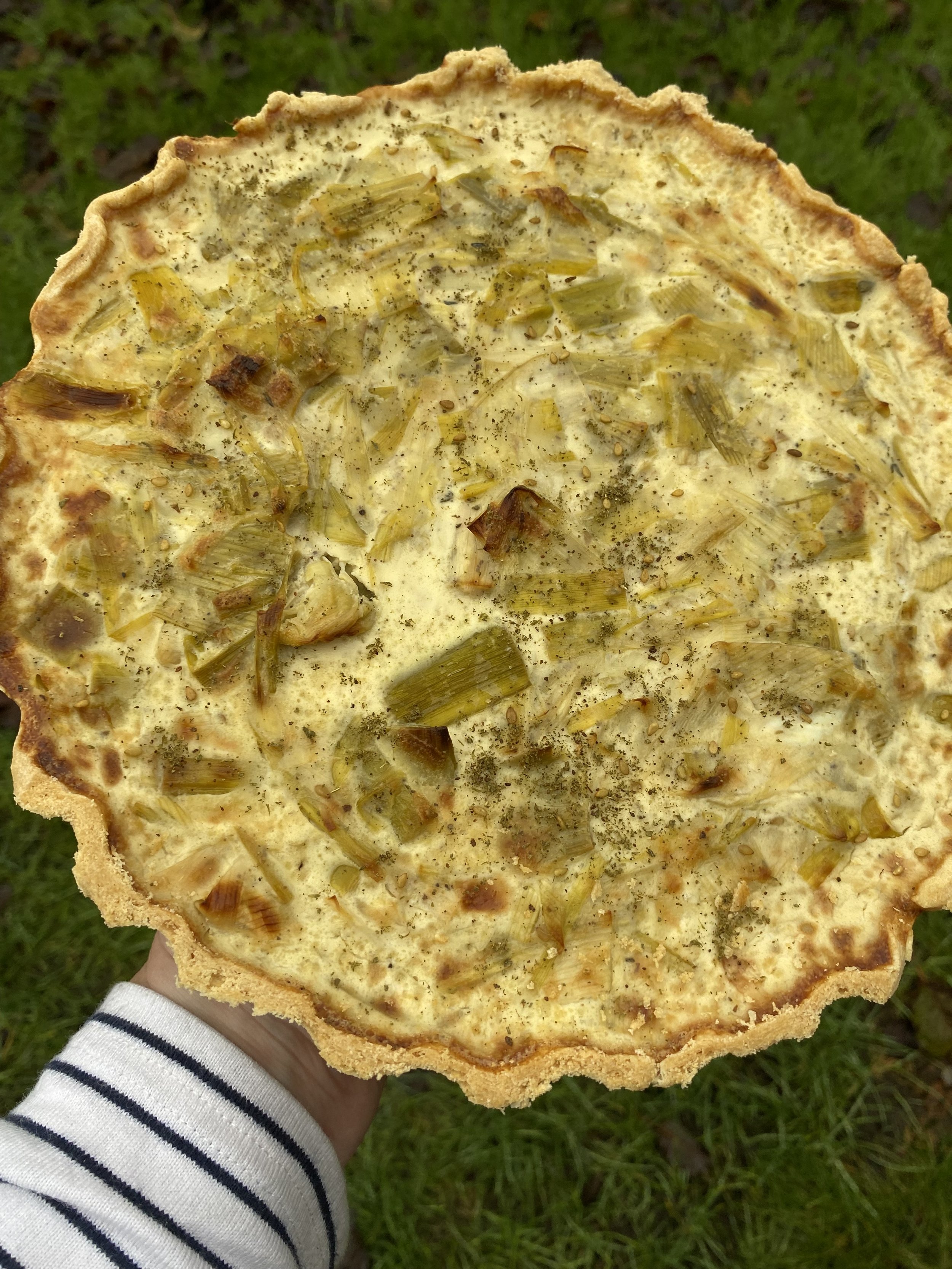 A person holding a homemade onion tart outside on green grass. The tart has a golden-brown crust and is topped with chopped cooked onions, black pepper, and other seasonings.