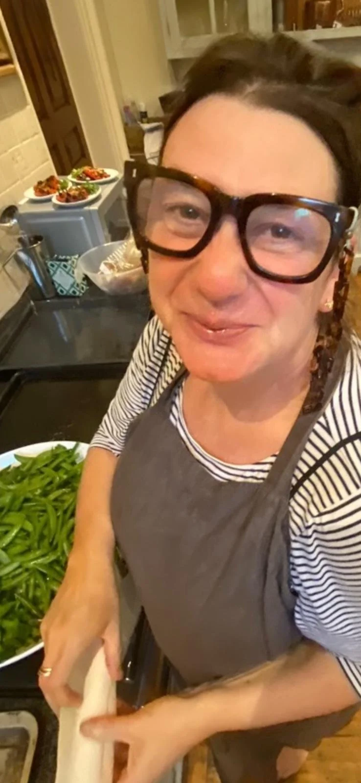 A woman with short hair, wearing large glasses and a striped shirt with an apron, is rolling dough in a kitchen. There are plates of cooked food on the counter in the background.