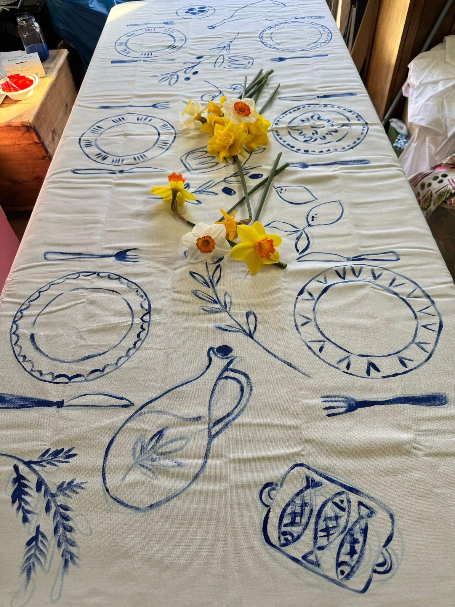 Supper club tablecloth with blue hand-painted plates, forks, spoons, and a pitcher design, decorated with a bouquet of yellow and white daffodils.