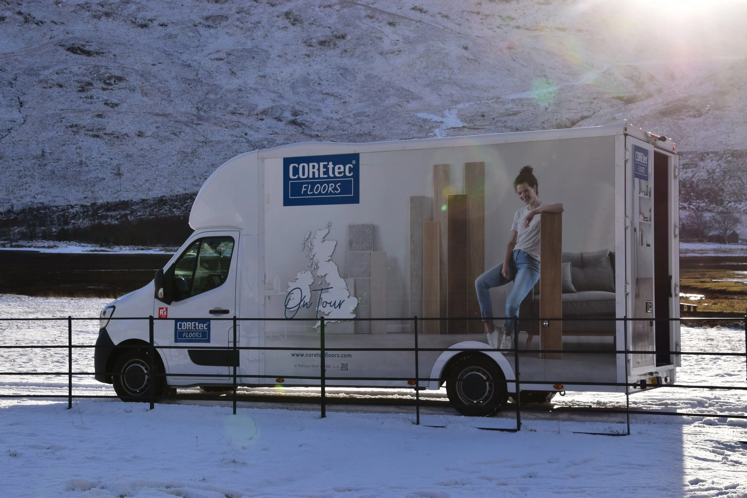 A white delivery truck with advertising for COREtec Floors parked behind a black fence in a snowy landscape with hills, trees, and a bright sky.