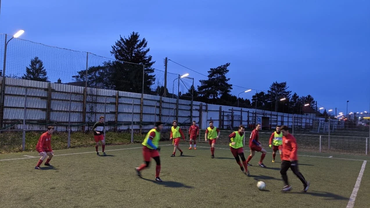 Jugendliche beim Fußballtraining auf einem Feld bei Dämmerung, einige in gelben Trainingswesten, andere in roten Trikots, auf einem Sportplatz mit Flutlicht.