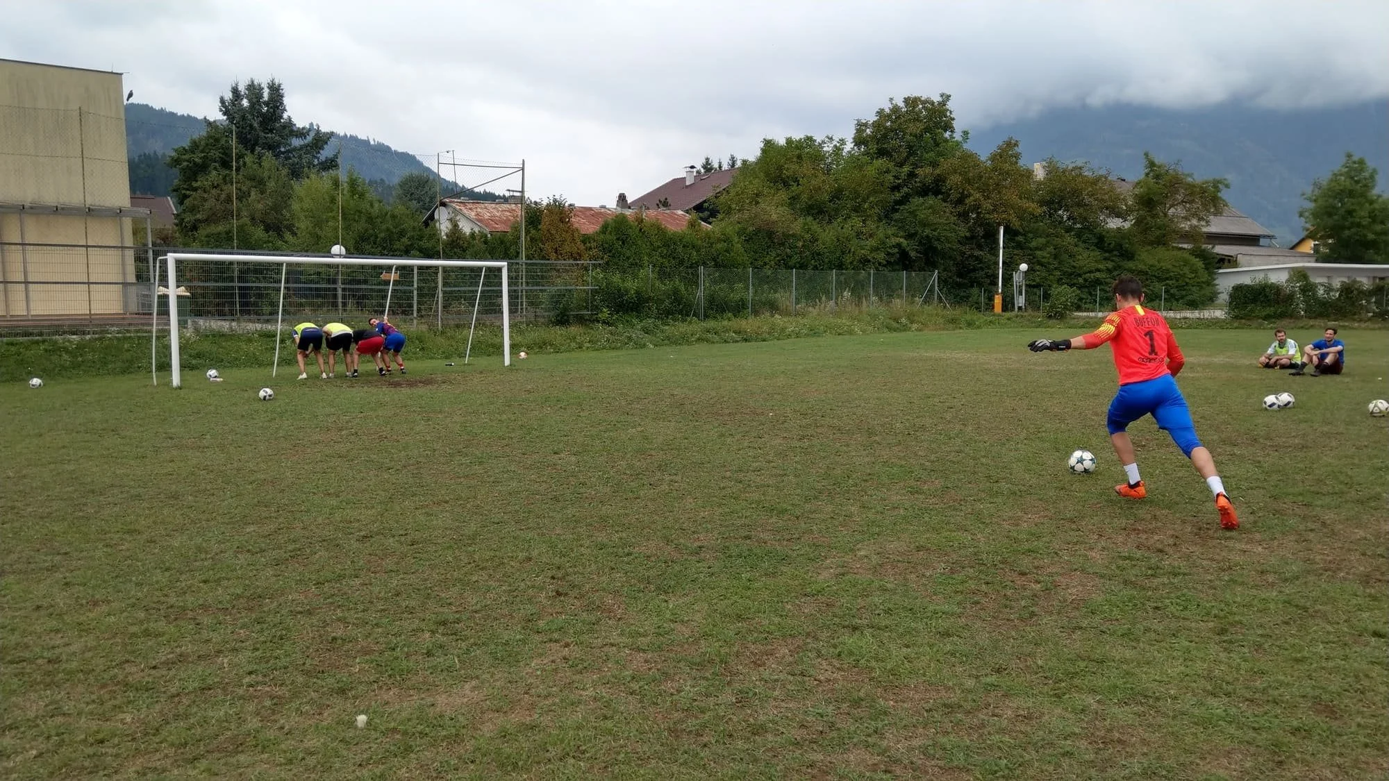 Fußballtraining mit Kindern auf einem Rasenfeld, einige Kinder üben Torwartfähigkeiten, während ein andere Spieler einen Freistoß vorbereitet, umgeben von mehreren Fußballbällen, bei bewölktem Himmel in einer ländlichen Umgebung.