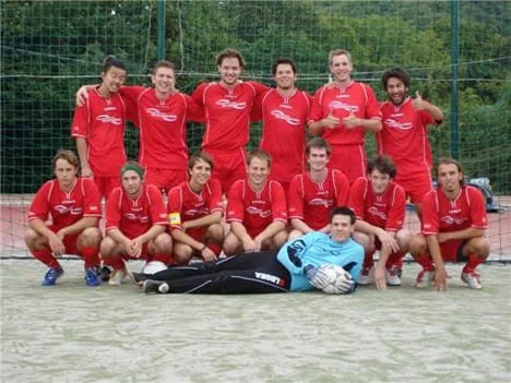 Fußballteam in roten Trikots posiert auf dem Spielfeld, mit Torwart in blauer Jacke liegend in der Mitte, draußen im Freien.