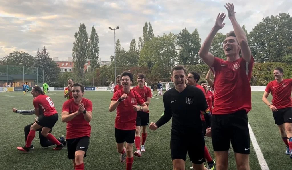 Glückliche Jugendfußballmannschaft feiert auf dem Spielfeld nach einem Spiel, einige jubeln, andere laufen lachend, im Hintergrund Bäume und Vereinsstände.