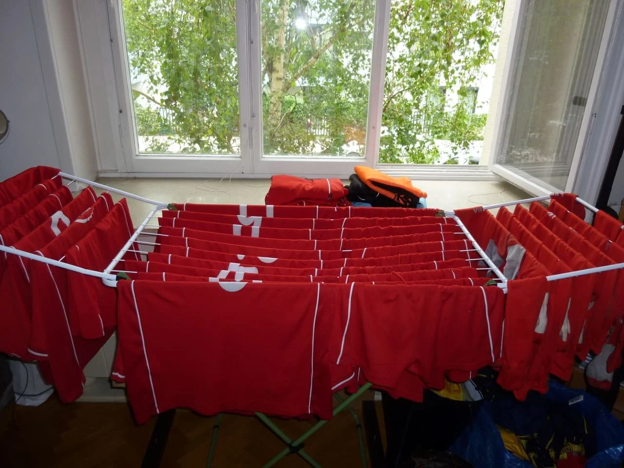 Bügelwäsche mit roten Trikots, hängen zum Trocknen auf einer Wäschestange vor einem Fenster mit grünen Bäumen draußen.