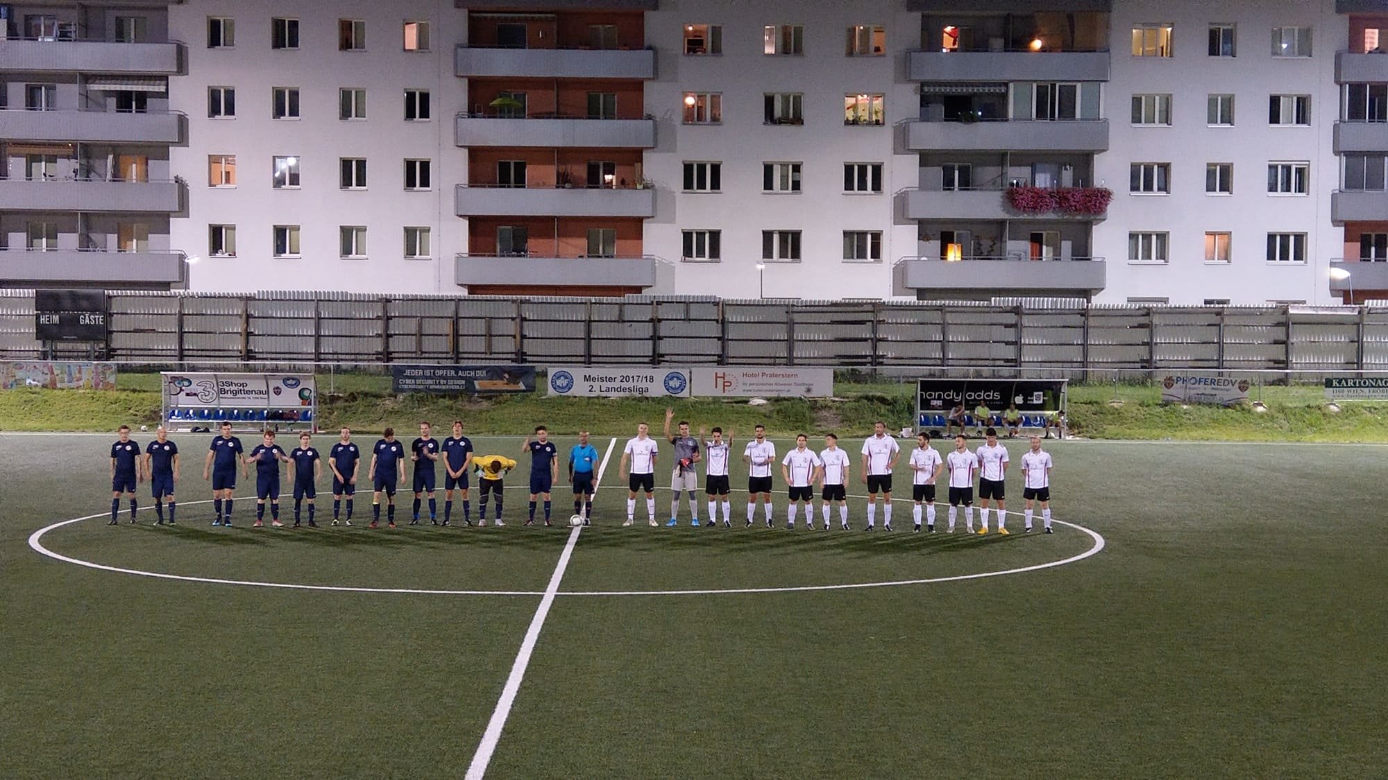 Fußballmannschaft steht in einer Reihe auf dem Spielfeld vor einem Wohngebäude, getragen in blauen und weißen Trikots, mit einem Schiedsrichter und einem Ball in der Mitte.