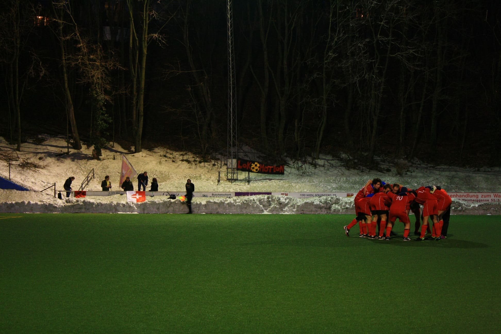 Fußballspieler in Rot feiern einen Torjubel auf einem grünen Rasenplatz, im Hintergrund Schneelandschaft und dunkler Wald bei Nacht.
