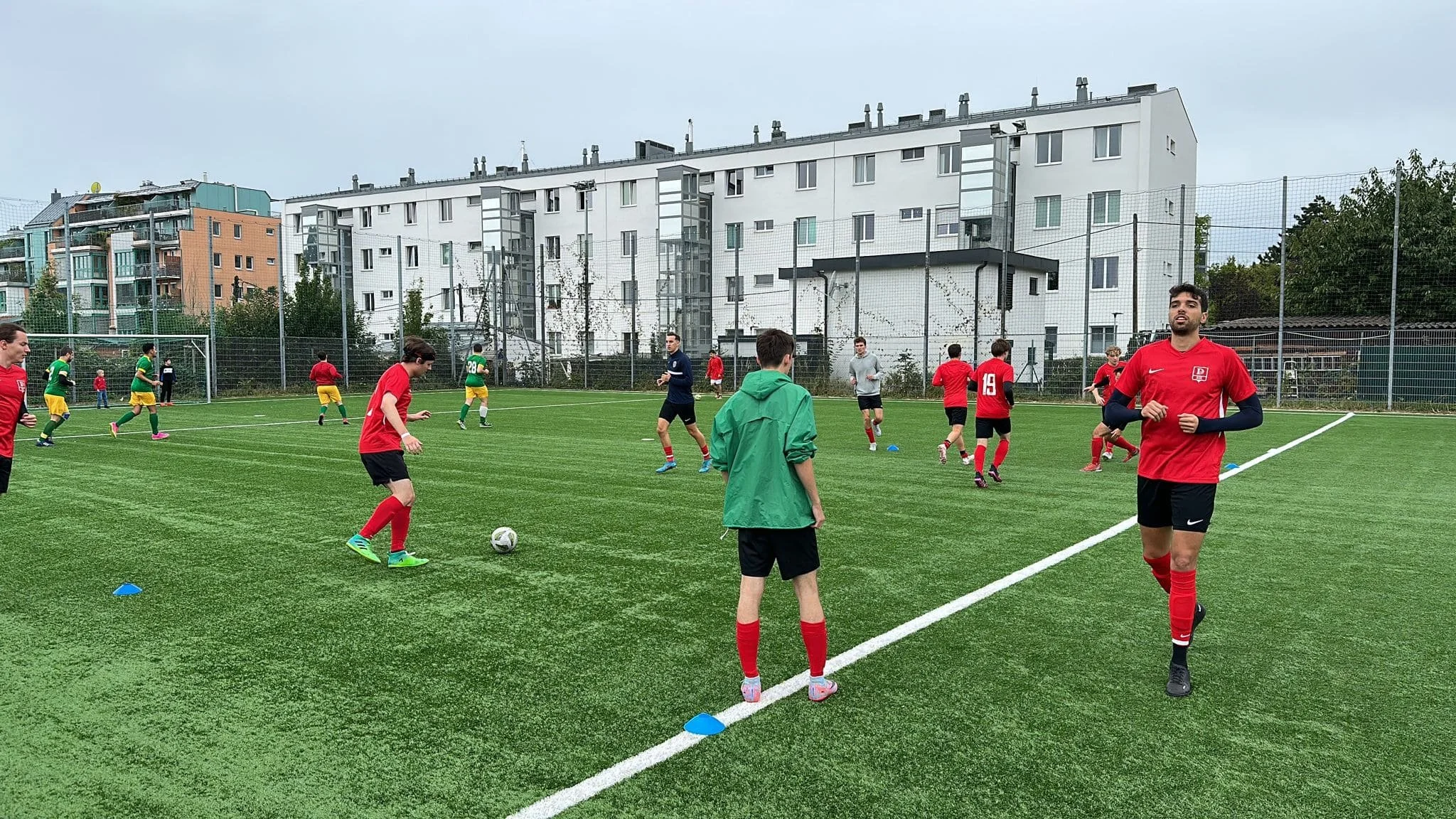Jugendliche beim Fußballtraining auf einem grünen Kunstrasenplatz, einige spielen, andere schauen zu, im Hintergrund Mehrfamilienhäuser, bewölkter Himmel.