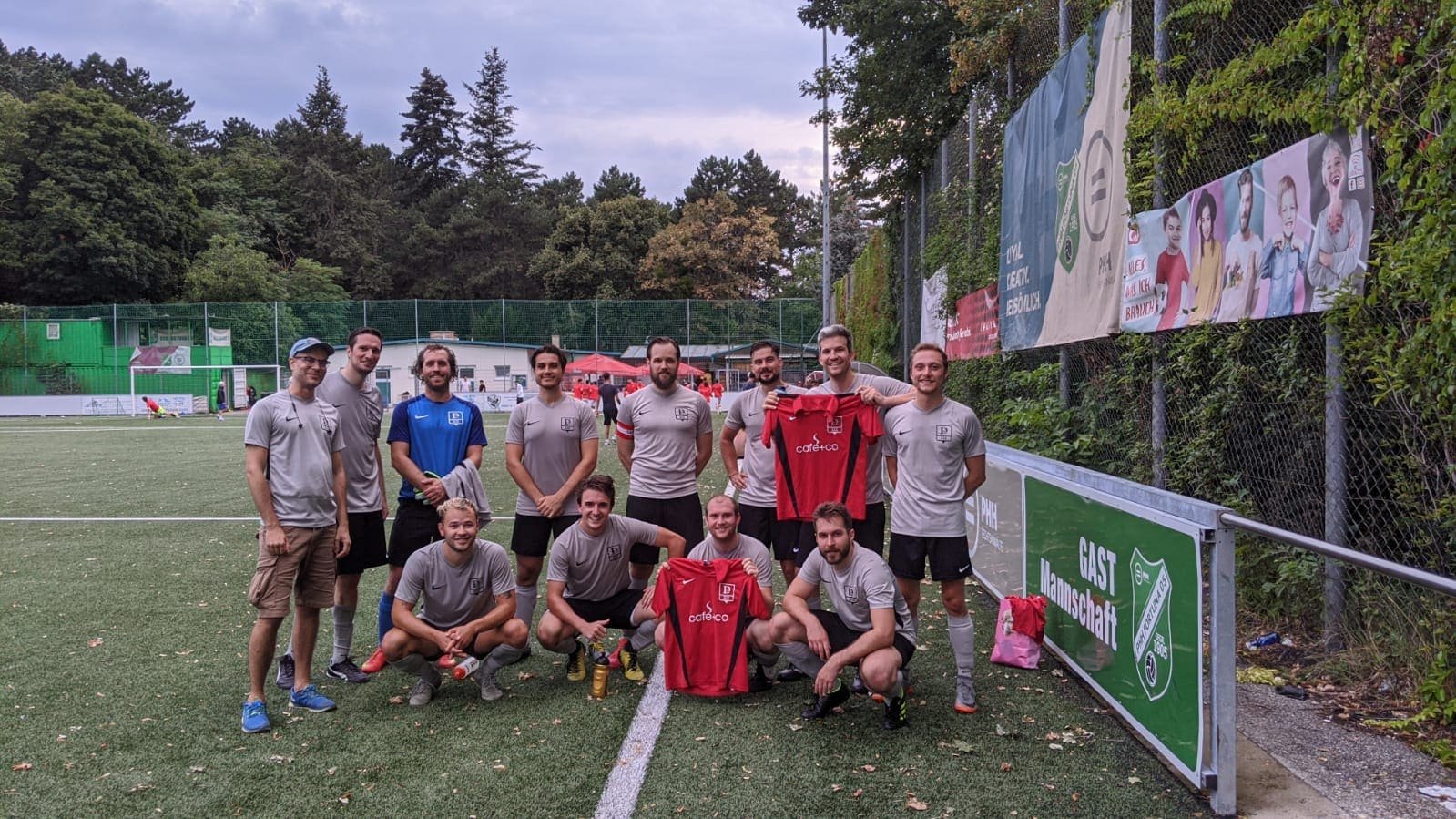 Eine Gruppe von elf Männern steht auf einem Fußballfeld, einige halten rote Fußballtrikots, im Hintergrund Bäume und Werbebanner.