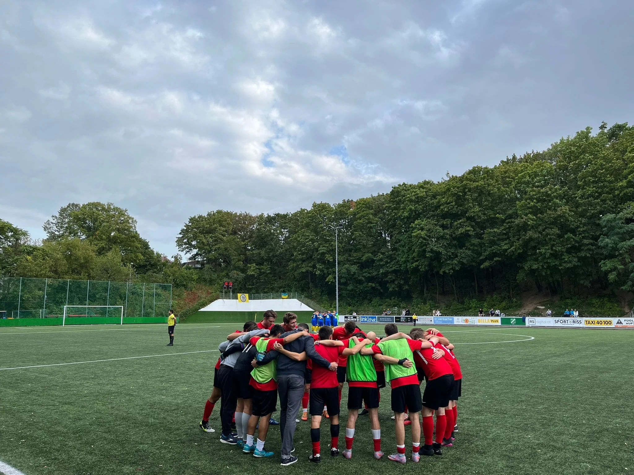 Fußballmannschaft in einer Gruppenumarmung auf einem Fußballfeld vor einem bewölkten Himmel, umgeben von Bäumen.