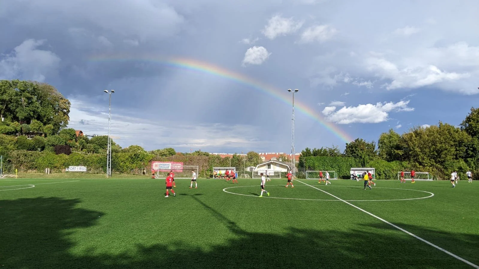 Fußballspiel auf einem grünen Rasenplatz mit einem Regenbogen im Himmel im Hintergrund.