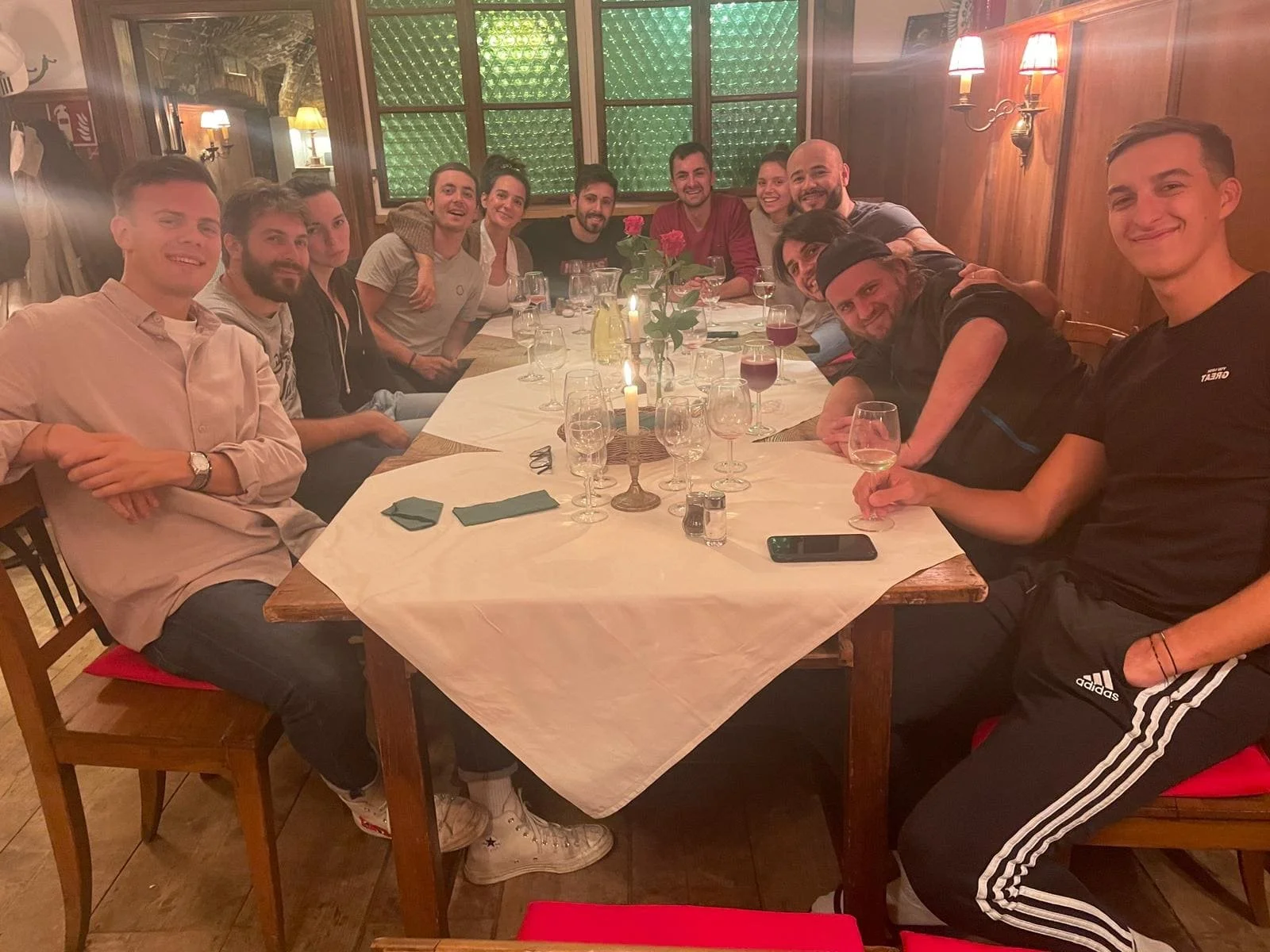 Gruppe von zwölf Menschen beim Abendessen in einem holzvertäfelten Raum, einige mit Weingläser, einige sitzen, einige stehen, Kerzen und eine Vase mit roten Rosen auf dem Tisch.