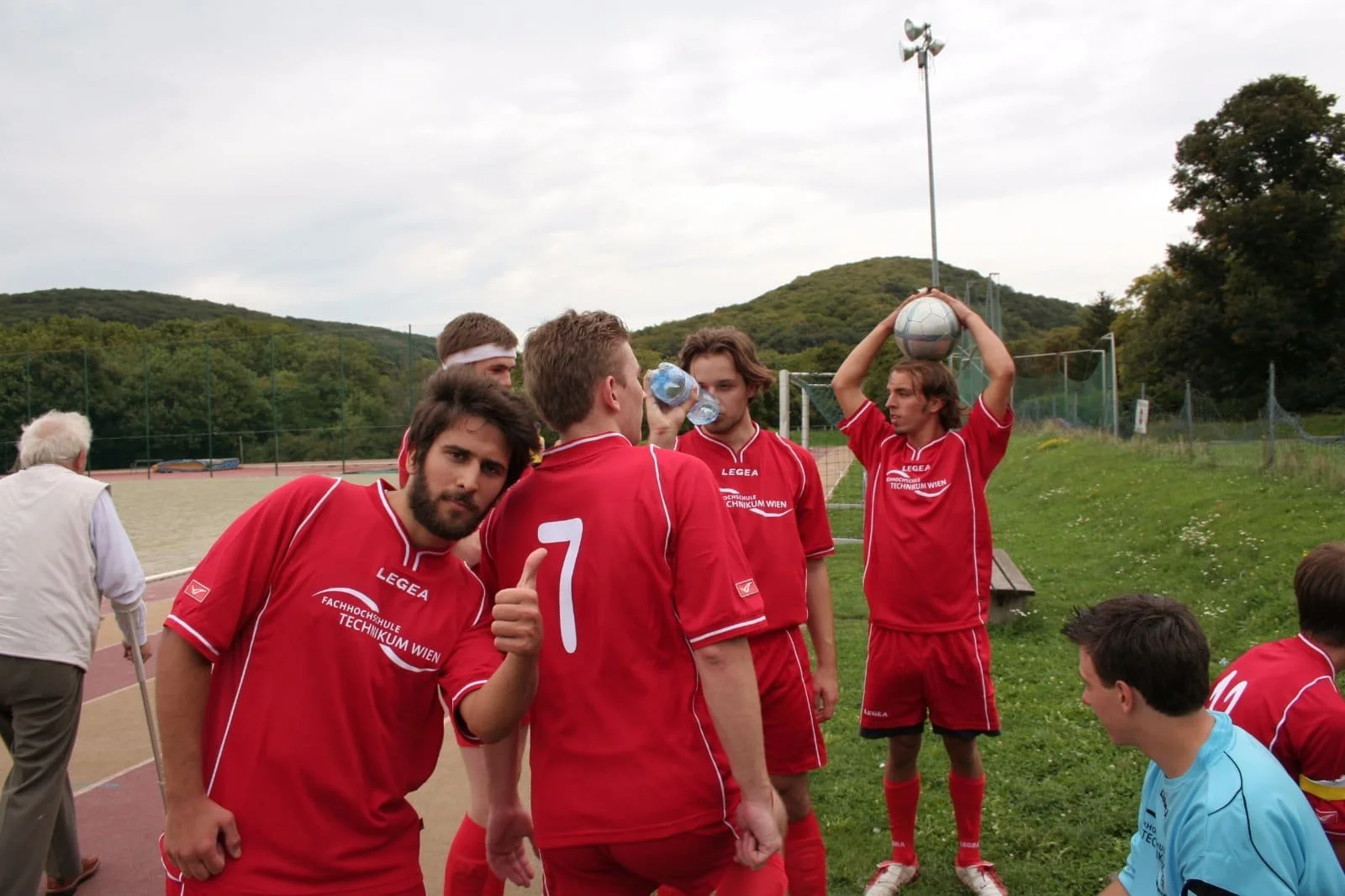 Fußballspieler in roter Uniform auf dem Spielfeld, ein Spieler macht einen Daumen hoch, ein anderer hält einen Wasserflasche, ein Spieler bereitet sich zum Einwerfen vor, ein Torwart sitzt neben den Spielern, im Hintergrund sind Bäume und Hügel zu se