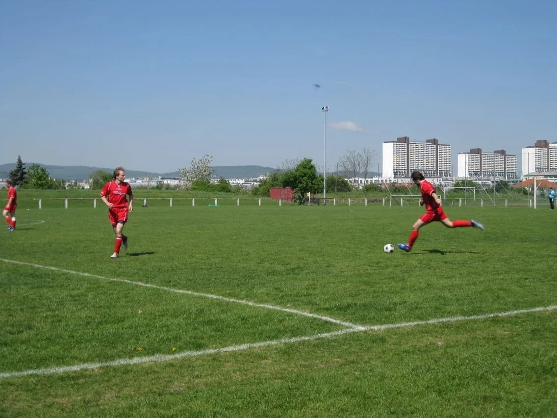 Fußballspieler in roten Trikots beim Spiel auf einem grünen Rasenplatz, mehrere Gebäude im Hintergrund, blauer Himmel.
