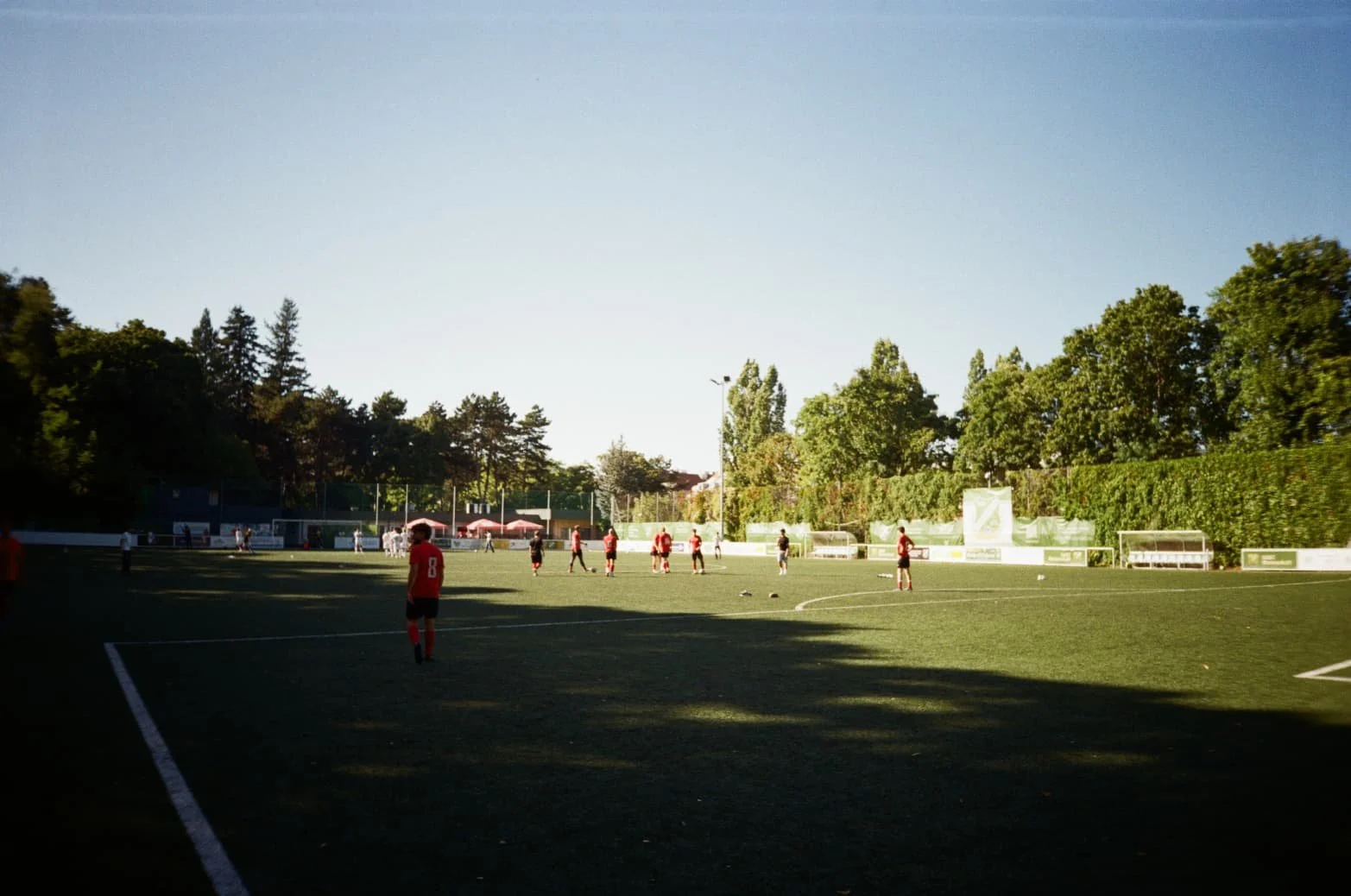 Fußballspiel auf einem Stadionplatz bei Sonnenuntergang mit Spielern in roten und schwarzen Trikots und Zuschauern im Hintergrund, umgeben von Bäumen.