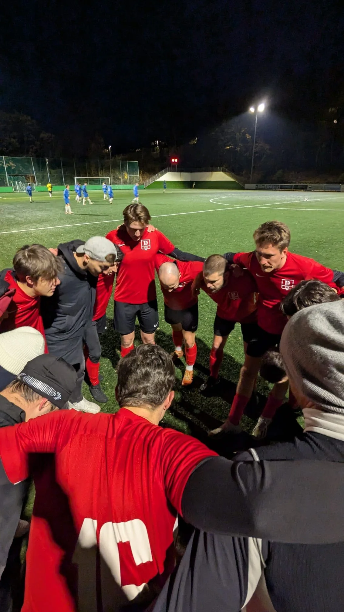 Fußballmannschaft in einer kreisförmigen Huddle auf einem beleuchteten Fußballfeld bei Nacht.