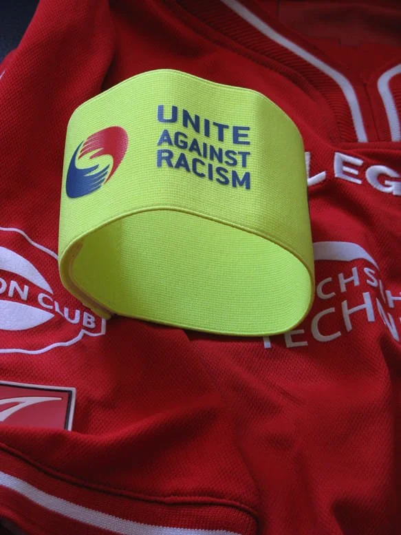 Gelbe Armband mit dem Text 'Unite Against Racism' und einem Logo, liegt auf einem roten Sporttrikot.