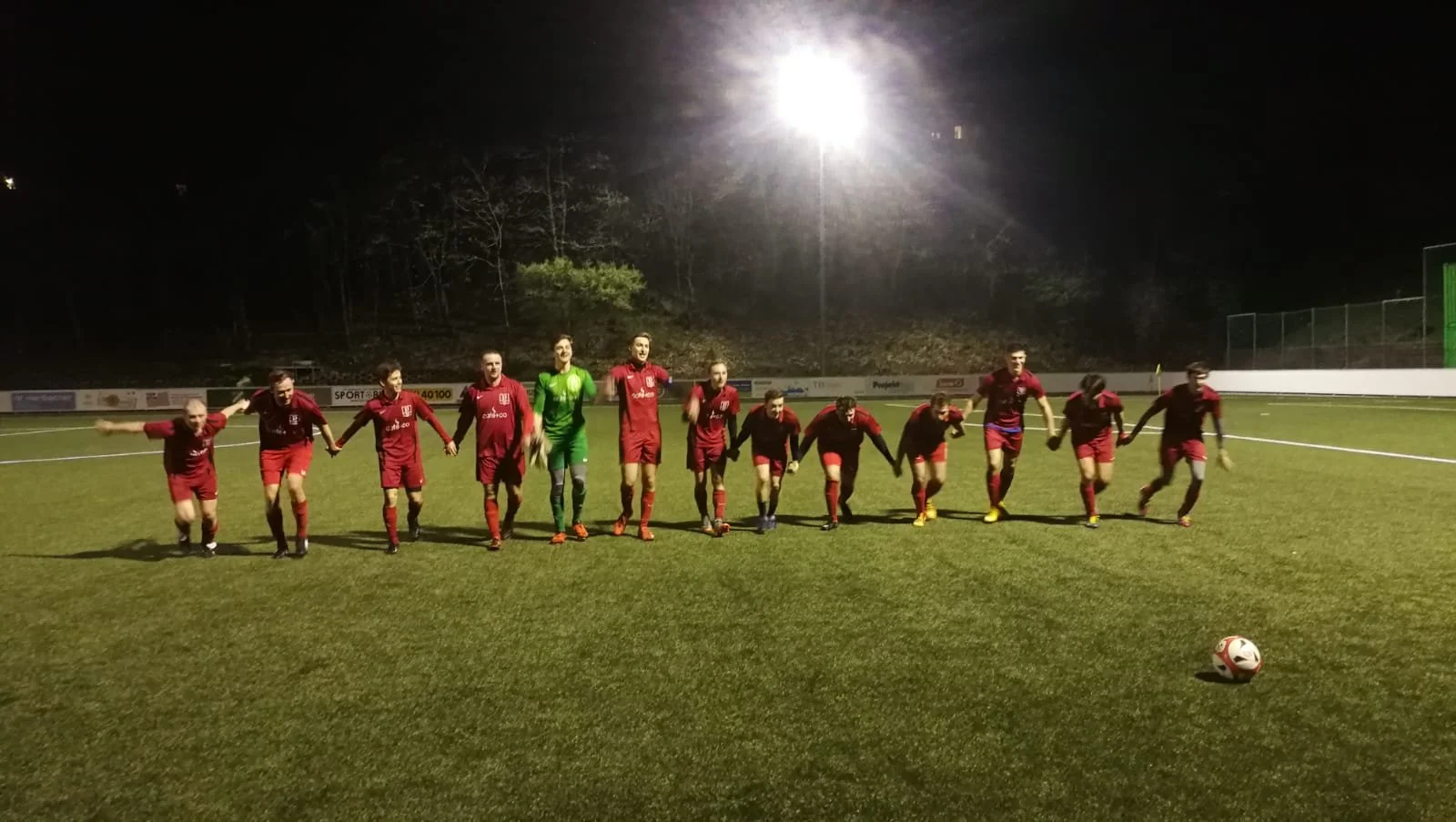 Fußballmannschaft in roten Trikots auf einem Spielfeld bei Nacht, Händchen haltend, mit einem Ball vor ihnen.