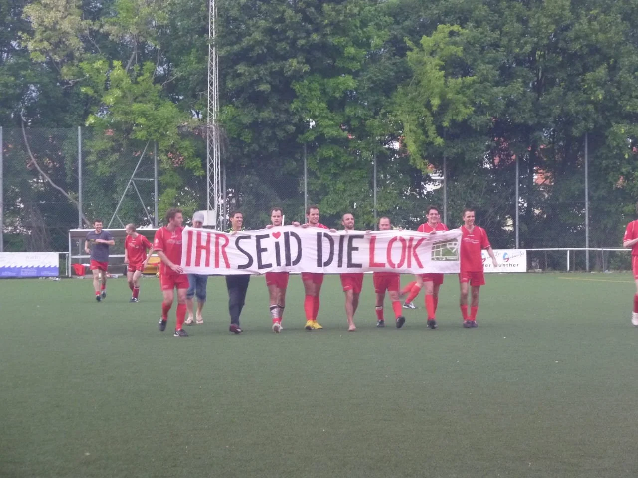 Fußballmannschaft in roten Trikots hält ein Banner mit der Aufschrift 'IHR SEID DIE LOK' auf einem Fußballplatz.