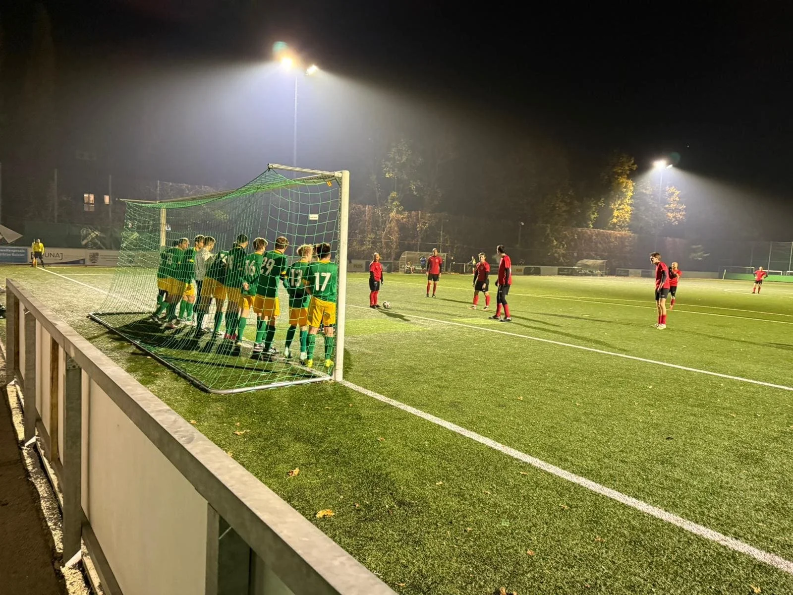 Fußballspiel bei Nacht auf einem künstlichen Rasenplatz, das Team in grünen Trikots und gelben Shorts steht in der Verteidigung, während die gegnerische Mannschaft in roten Trikots in der Nähe des Strafraums positioniert ist.