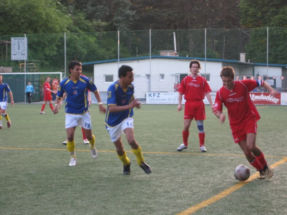 Fußballspiel auf einem Kunstrasenplatz mit Spielern in blauen und roten Trikots, einige laufen hinter einem Spieler in roten Trikots, der den Ball schießt.