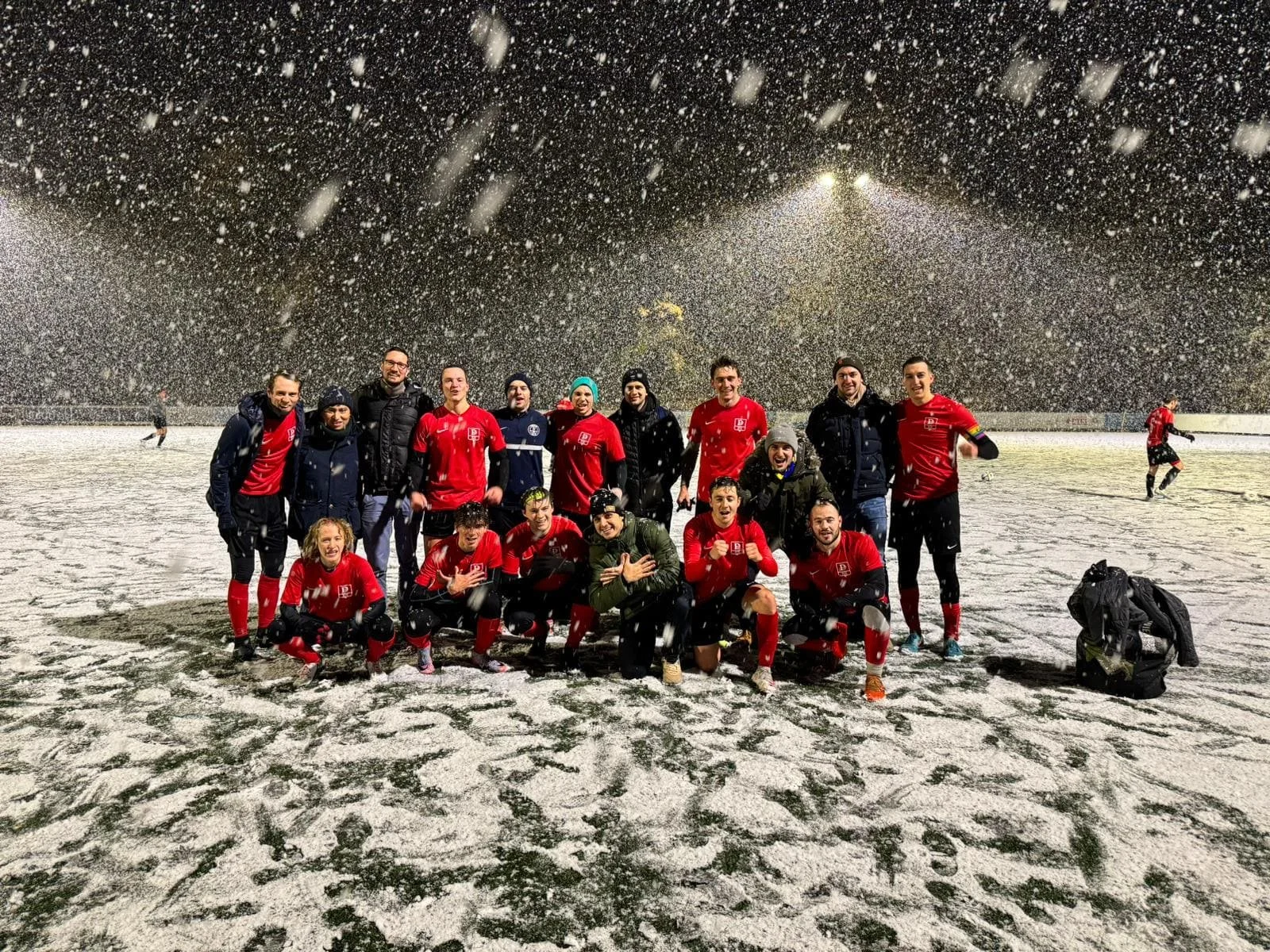 Gruppe von Fußballspielern bei Schneefall nach einem Spiel auf dem Spielfeld, einige jubeln, halten den Daumen hoch, in Sportkleidung, nächtlicher Himmel, Stadionbeleuchtung