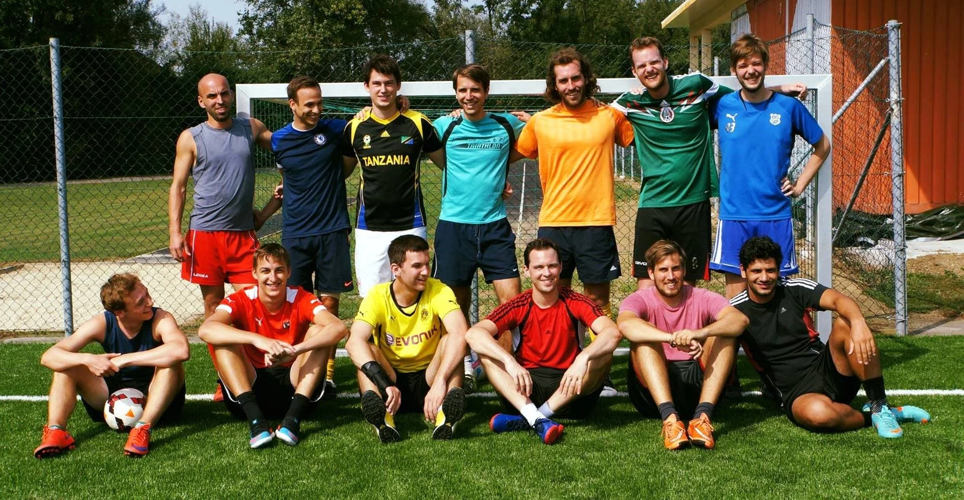 Gruppe von jungen Männern auf einem Fußballplatz, posieren vor einem Tor, einige sitzen auf dem Boden, andere stehen hinter ihnen, alle tragen Sportkleidung.