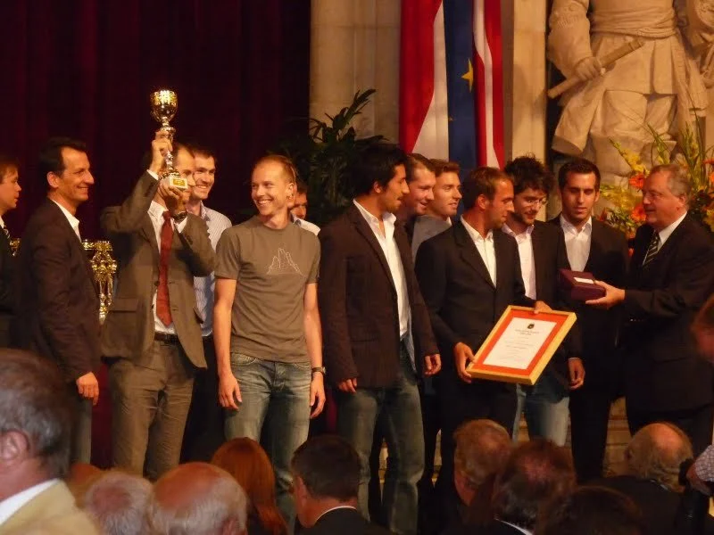 Auszeichnung eines Teams bei einer Zeremonie, ein Mitglied hält einen Pokal, andere halten eine Urkunde, lautes Applaudieren, im Hintergrund Nationalflaggen und eine Skulptur.