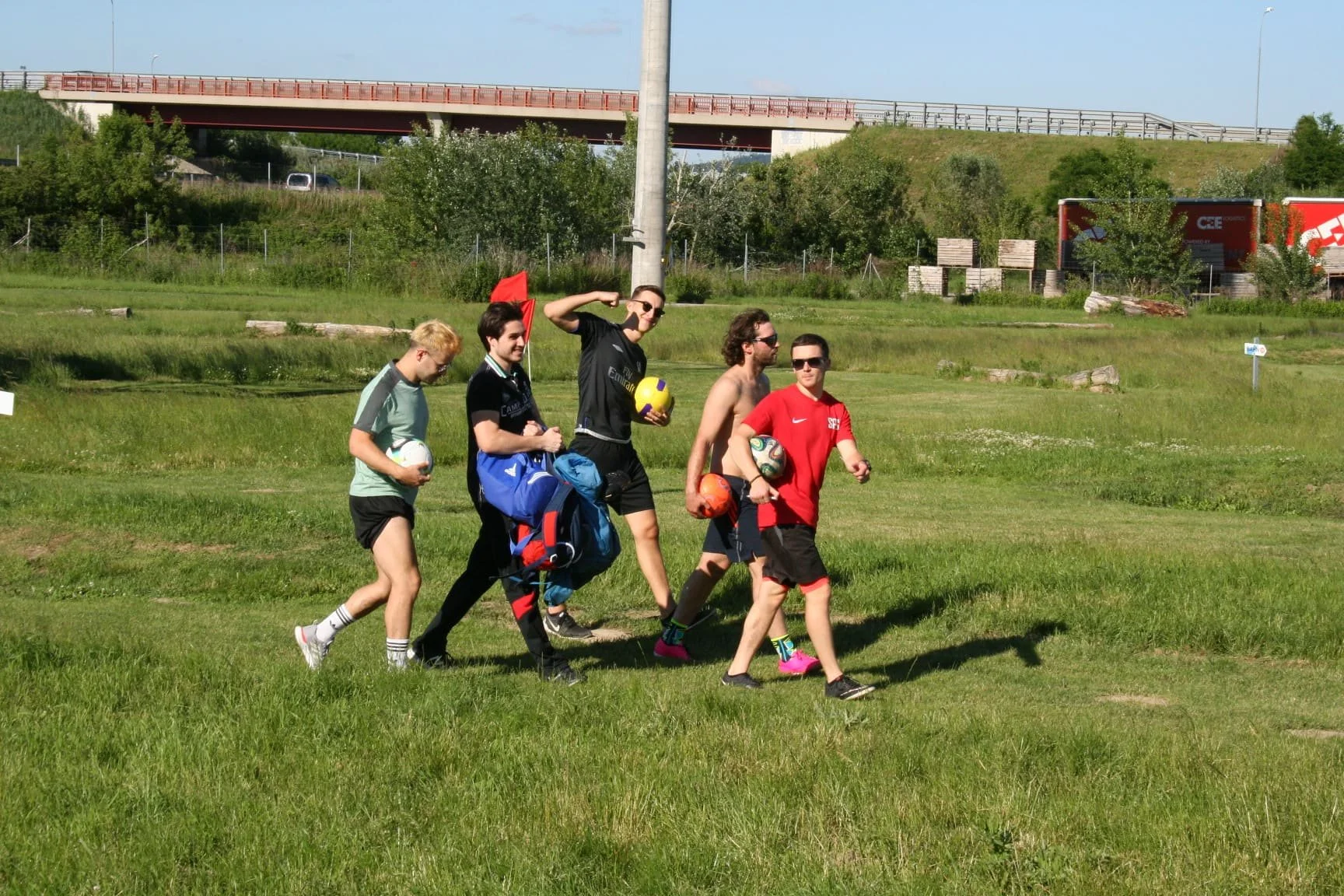 Fünf junge Männer gehen auf einem grünen Rasen, einige halten Fußball».}}Skład artykułu:  Pięciu młodych mężczyzn na pastwisku, niektórzy trzymają piłkę nożną. Oświetlenie: słoneczne, pogoda jest jasna i ciepła. Tło: trawa, drzewa, podmostie i samoch