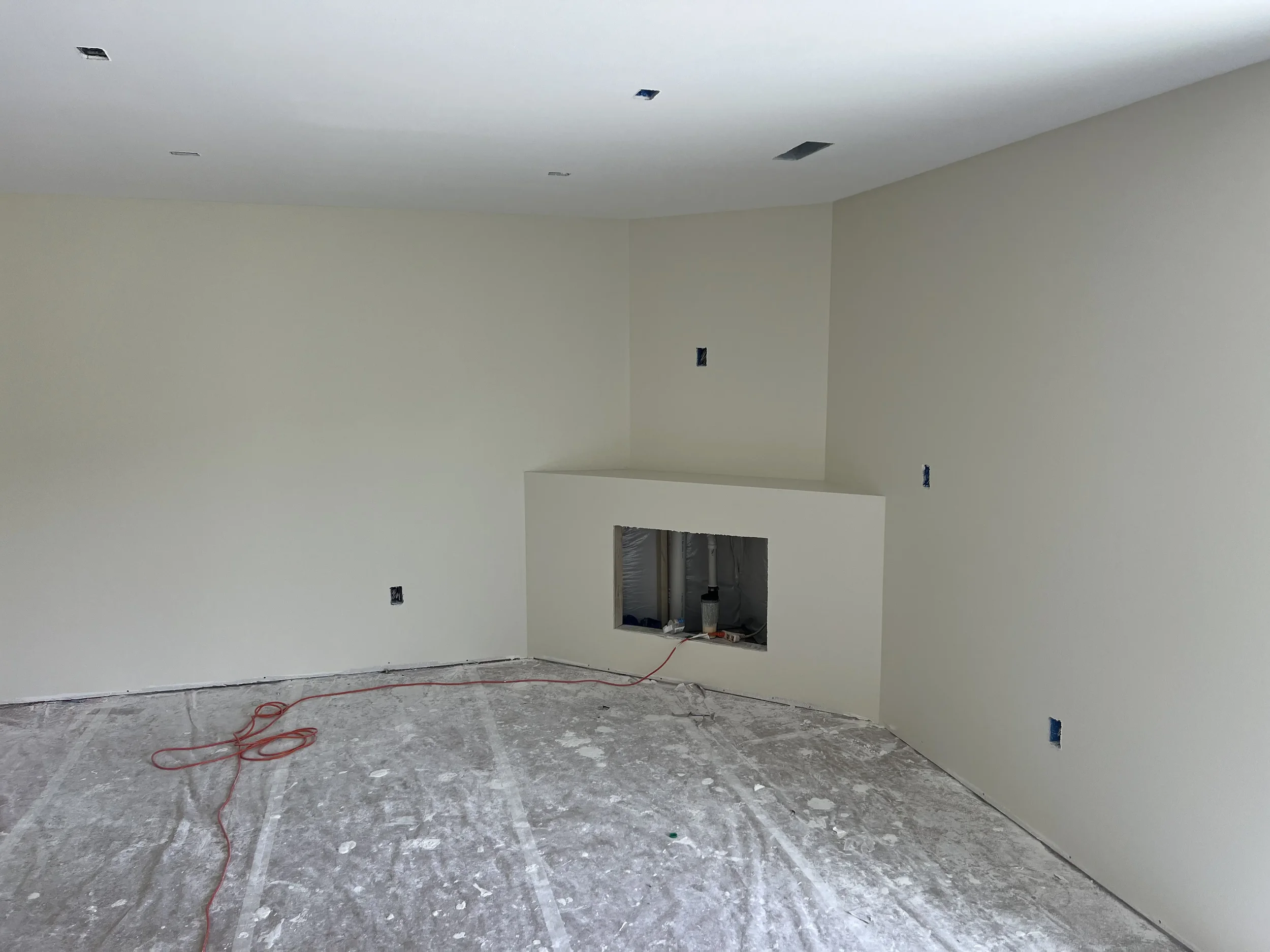 Empty room under construction with white walls, unfinished ceiling with multiple recessed lighting fixtures, and a floor covered in construction dust. There is a cutout in the wall indicating plumbing work, and several electrical outlets and wires vi