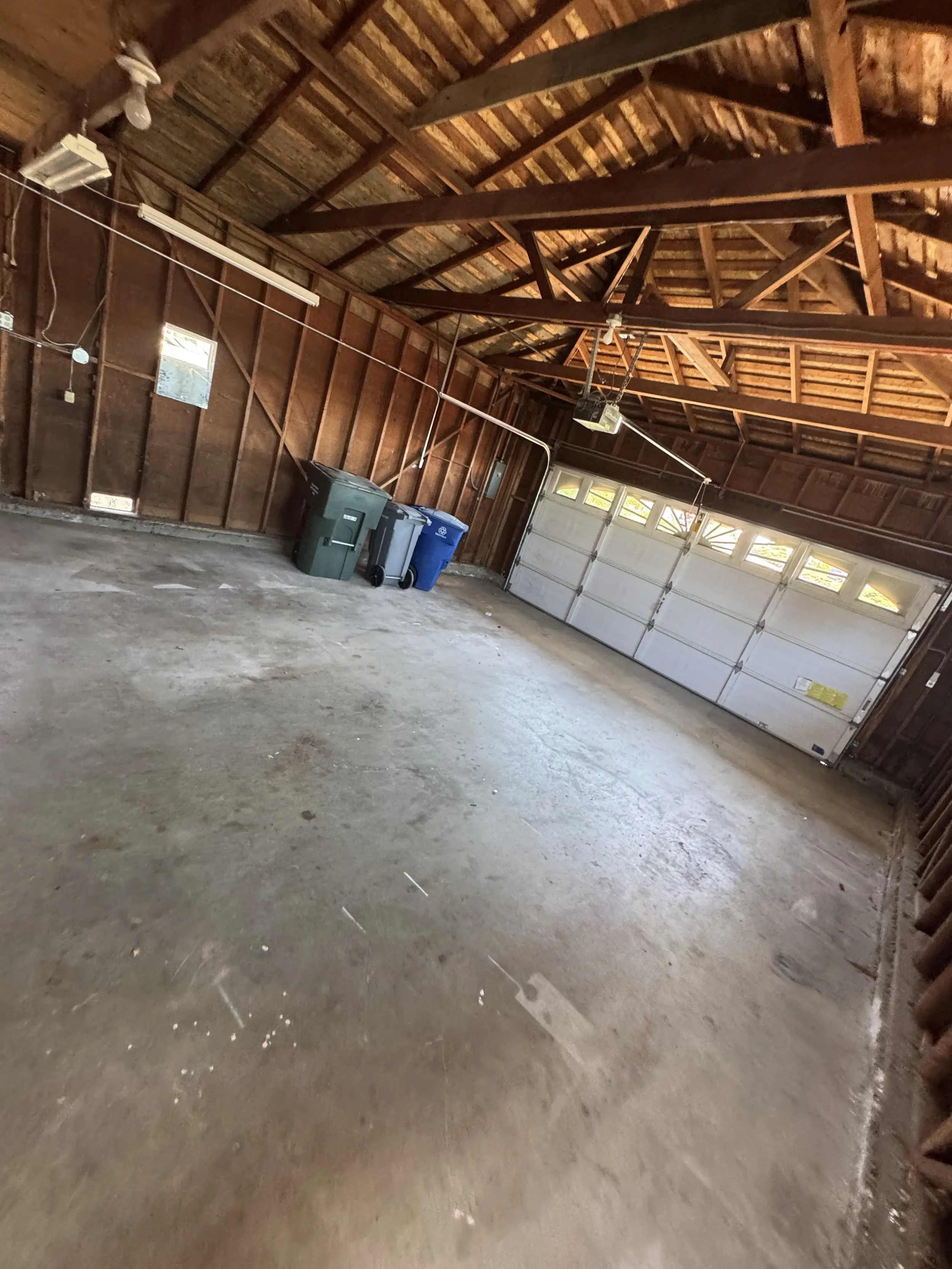 Empty garage with a concrete floor, wooden walls, and a wooden ceiling with exposed beams. There are four garbage bins near a closed garage door with windows at the top.
