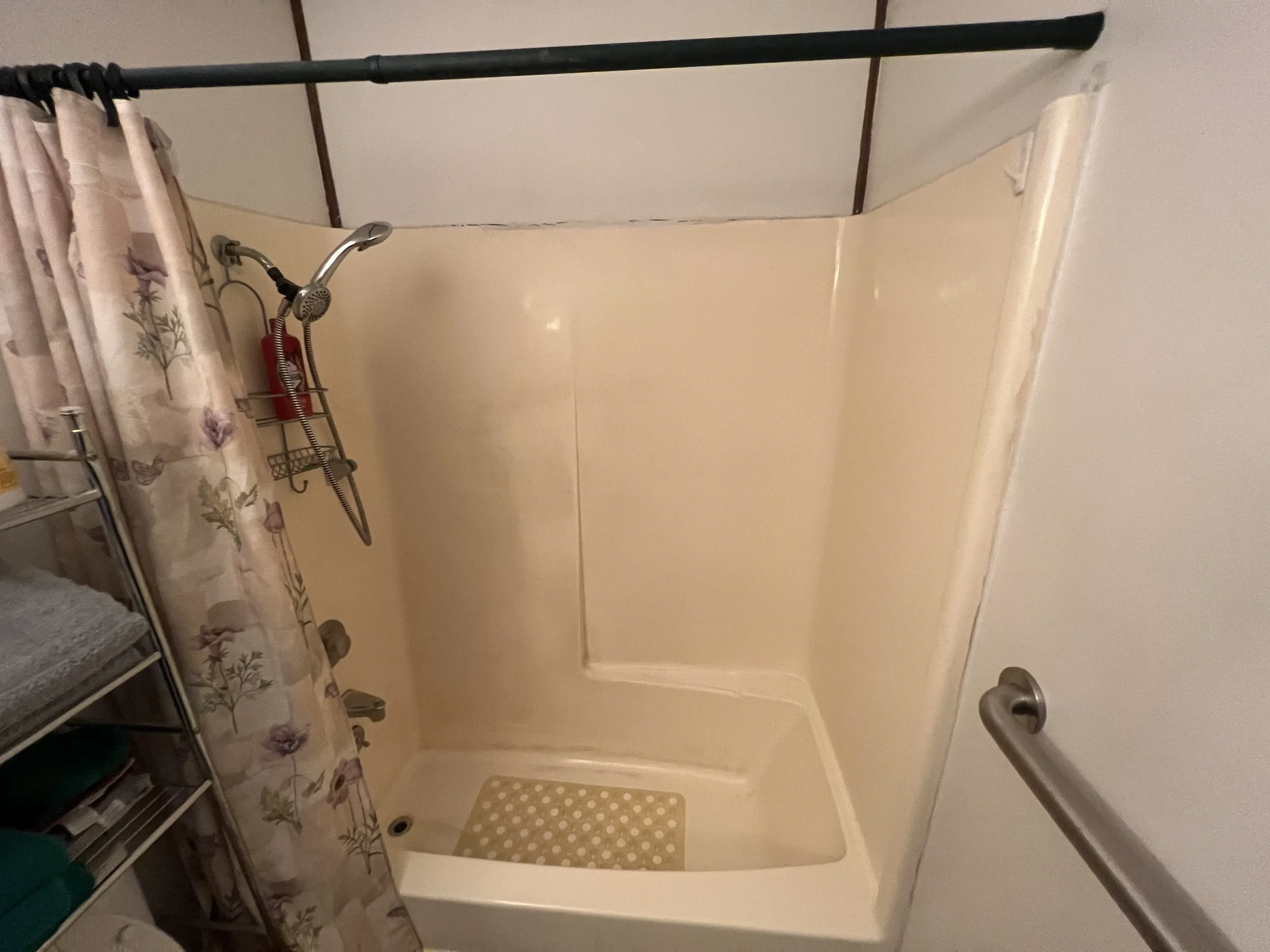 A shower with a curved shower wall, a handheld showerhead, a fire extinguisher, a floral shower curtain, and a chair with towels.