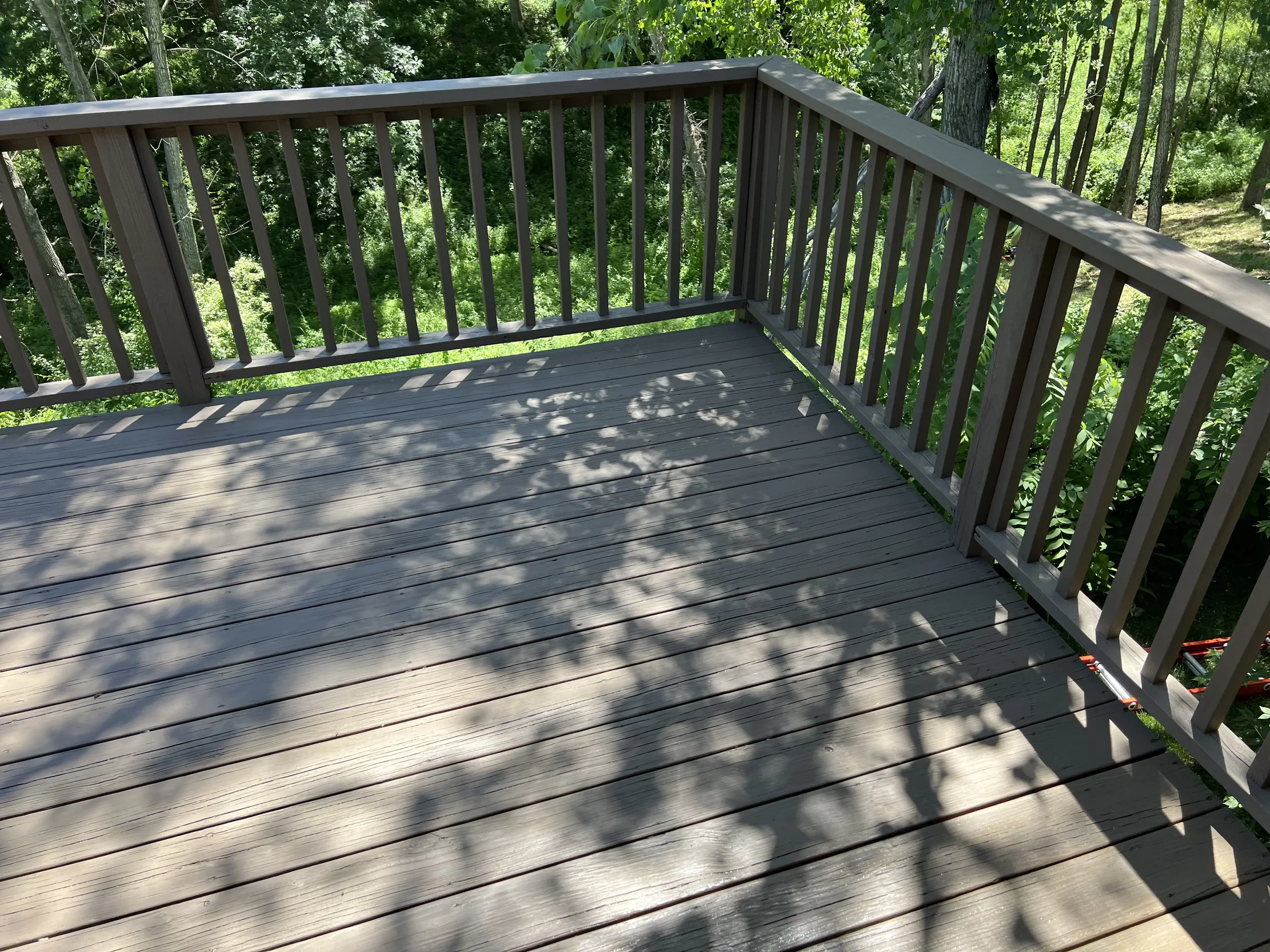 View of a wooden balcony with railing, cast shadows of tree branches and leaves, surrounded by green trees and bushes.