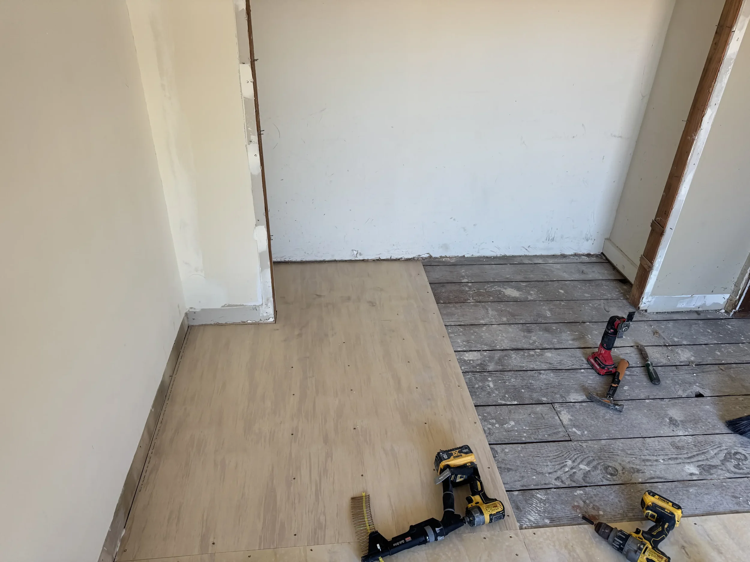 Interior room under renovation showing new wooden flooring being installed, with tools like a cordless drill, hammer, and screwdriver on the subfloor.