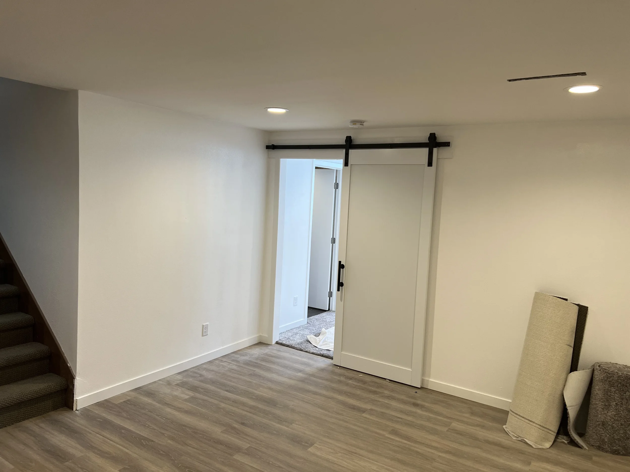 Empty room with white walls, wood flooring, a sliding barn door, and some rolled-up carpets leaning against the wall.