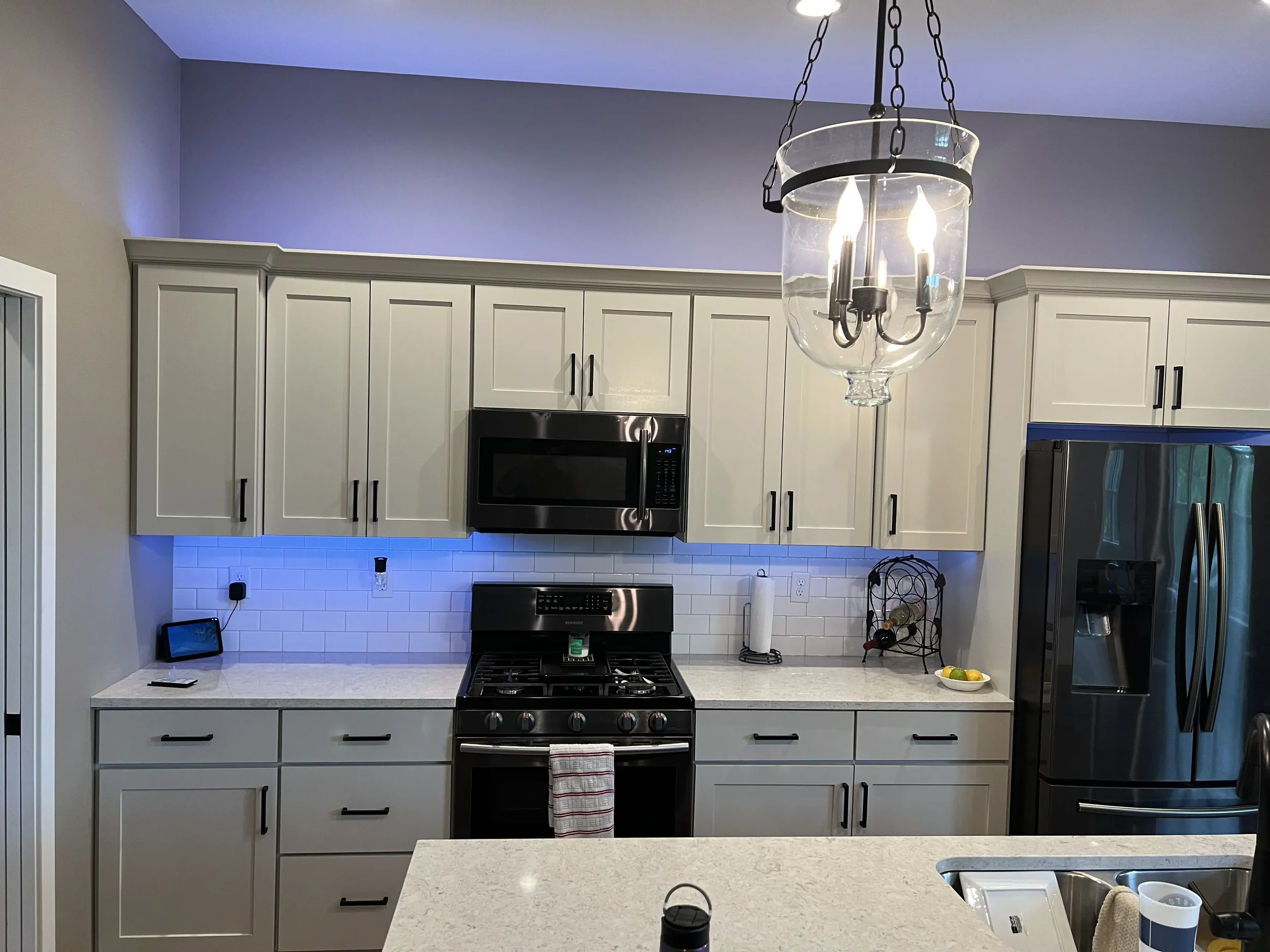 Modern kitchen with white cabinets, stainless steel appliances, a black oven and microwave, a white subway tile backsplash, a white countertop with a paper towel holder, a wine rack, a bowl with lemons and limes, a refrigerator with a water and ice d