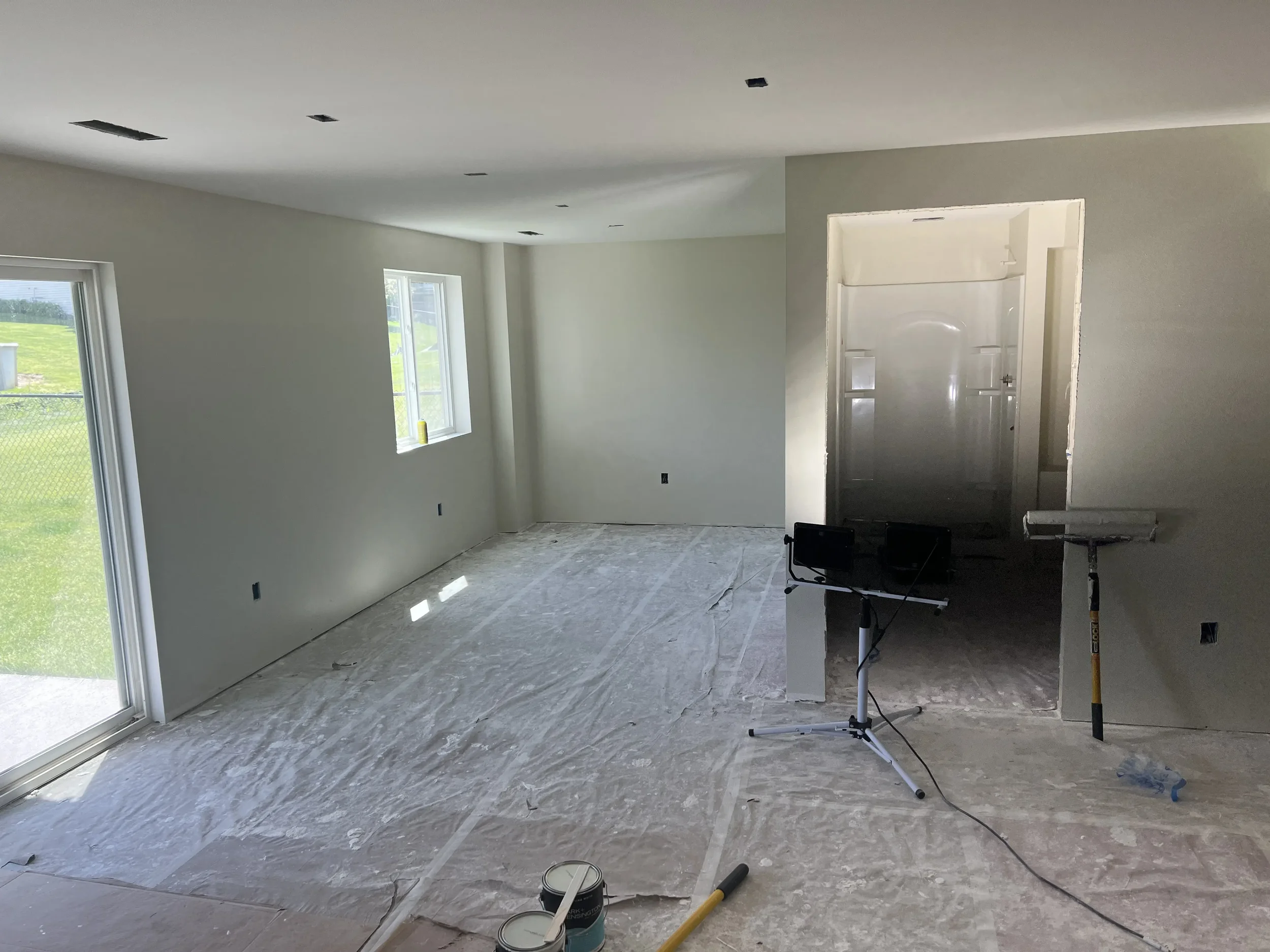 Interior of a house under construction with unfinished walls and ceiling, paint cans and construction tools on the floor, and a doorway leading to a metal room or space.