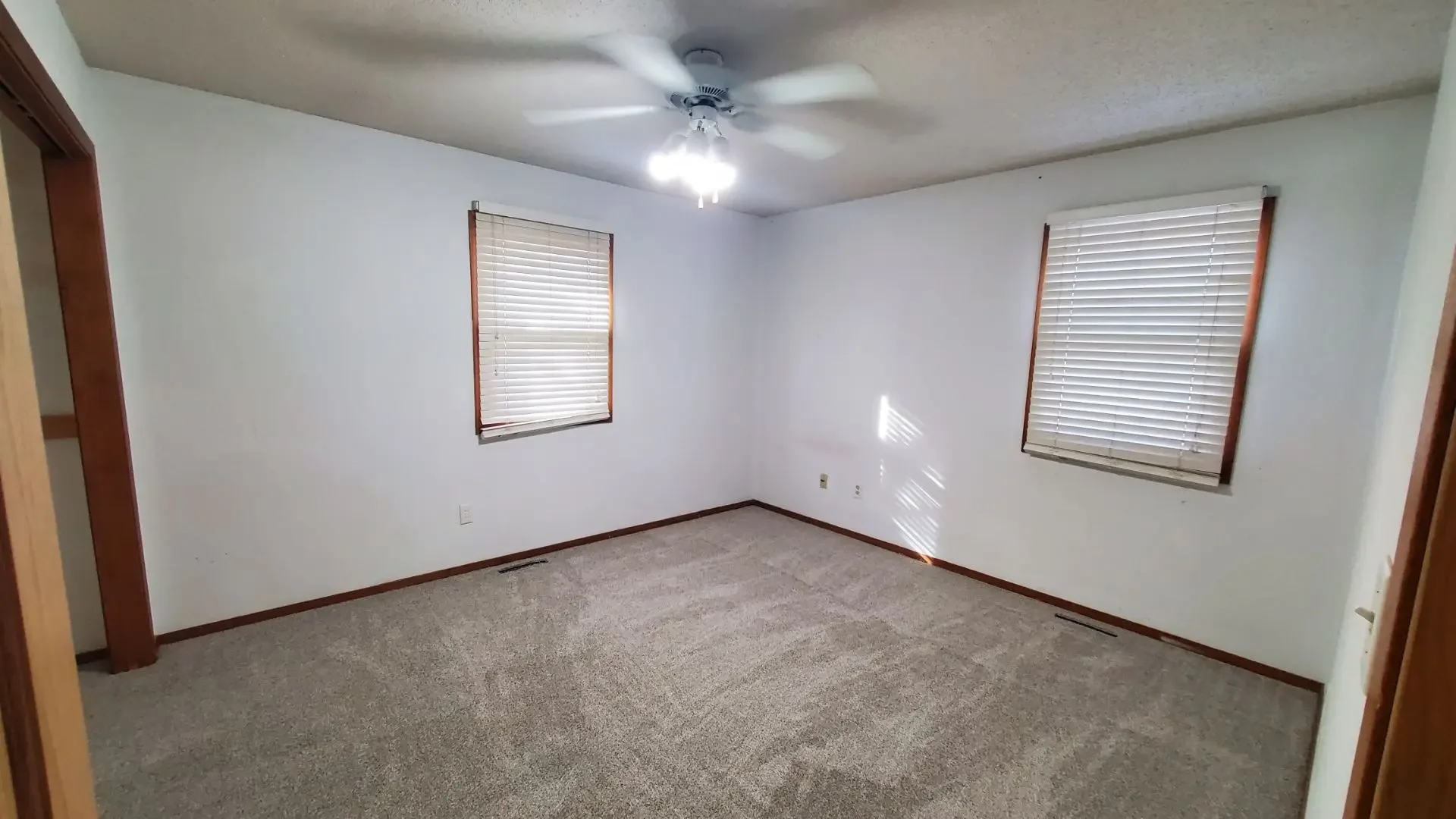 Empty room with beige carpet, white walls, two windows with blinds, a ceiling fan with lights, and wooden trim.