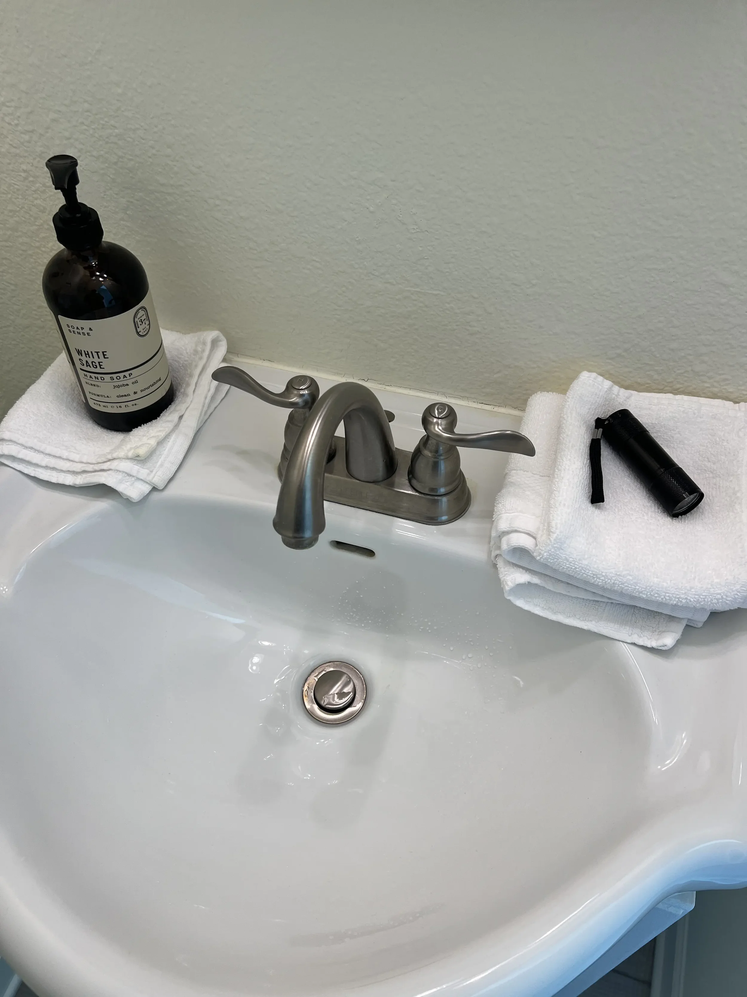 Bathroom sink with a bottle of hand soap, a folded towel, a flashlight, and a washcloth on opposite sides.